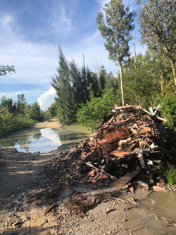 A pile of construction debris, including wooden planks and bricks, is situated beside a muddy path lined by trees and vegetation. In the background, a clear sky with scattered clouds can be seen reflecting in a small pool of water. The scene captures an area that contrasts natural greenery with the remnants of human activity. Bird Basin Park mountain bike trail.