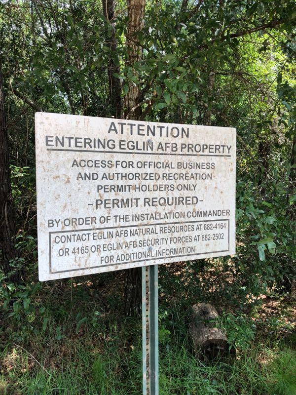 Sign indicating access restrictions for Eglin Air Force Base properties, stating that entry is allowed for official business and authorized recreation only with a required permit. The sign includes contact information for Eglin AFB Natural Resources and Security Forces for further inquiries. Surrounding foliage is visible, suggesting a wooded area. Rocky Creek Trails mountain bike trail.