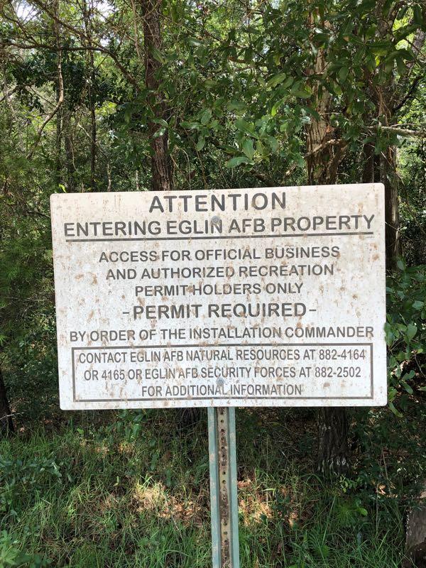 Sign indicating restricted access to Eglin Air Force Base property, stating that permits are required for official business and authorized recreation. Contact information for Eglin AFB Natural Resources and Security Forces is provided. Surrounded by greenery. Rocky Creek Trails mountain bike trail.