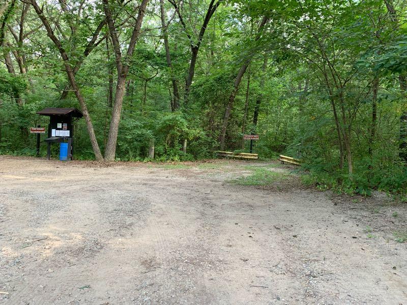 A gravel path leading into a wooded area, with a small kiosk and blue trash can on the left. Two wooden benches are visible nearby, along with signs indicating the trails ahead. Lush green trees surround the entrance, creating a natural, serene environment. Ortonville trail mountain bike trail.