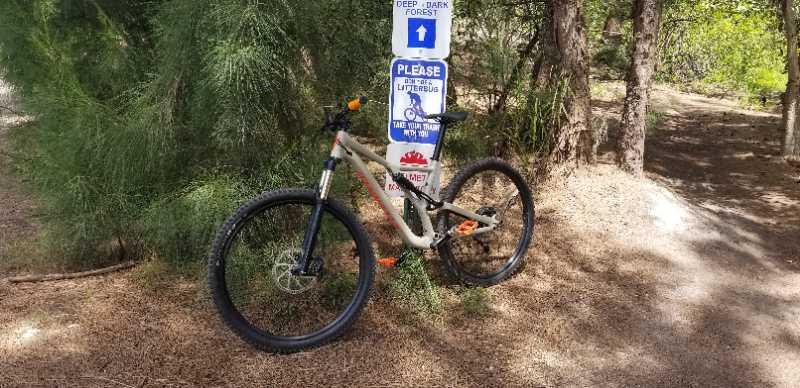 A mountain bike resting against a sign in a forested area, with a warning about deep and dark forest paths. The background features trees and a dirt trail, creating a natural outdoor setting. Markham Park mountain bike trail.