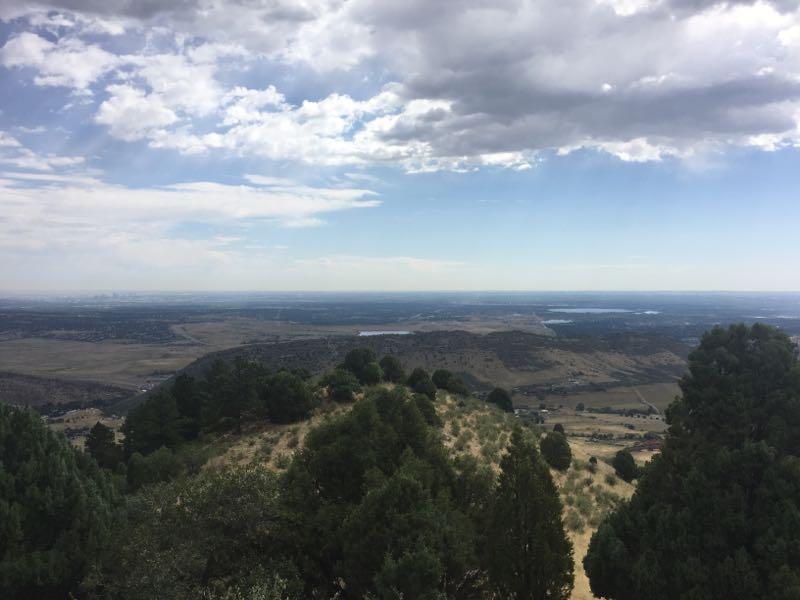 A panoramic view of rolling hills and valleys under a partially cloudy sky. The foreground features patches of green trees, while the horizon stretches out to show distant land features, including water in the background. The landscape appears expansive and serene, suggesting a natural setting. Mount Falcon Park mountain bike trail.