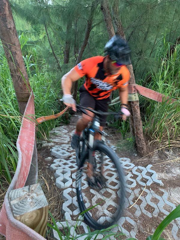 A cyclist riding a mountain bike on a narrow, winding trail surrounded by tall grass and trees. The rider is wearing an orange and black jersey and a helmet, and the motion blur suggests high speed as they navigate over textured paving stones. Virginia Key North Point mountain bike trail.