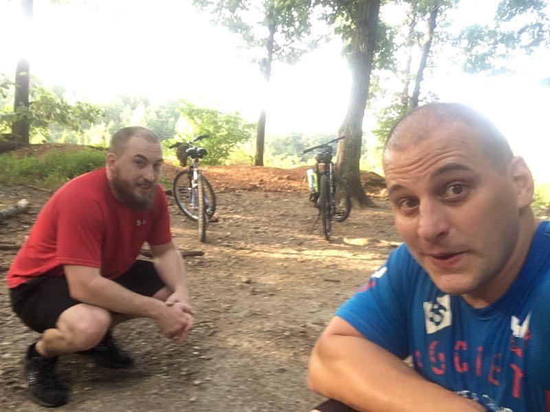 Two men are posing for a selfie in a forested area, smiling at the camera. One man is crouched down on the left, wearing a red shirt and black shorts, while the other is sitting in the foreground on the right, wearing a blue shirt. Two bicycles are parked in the background among trees, and the setting appears to be a recreational area. Enterprise South mountain bike trail.