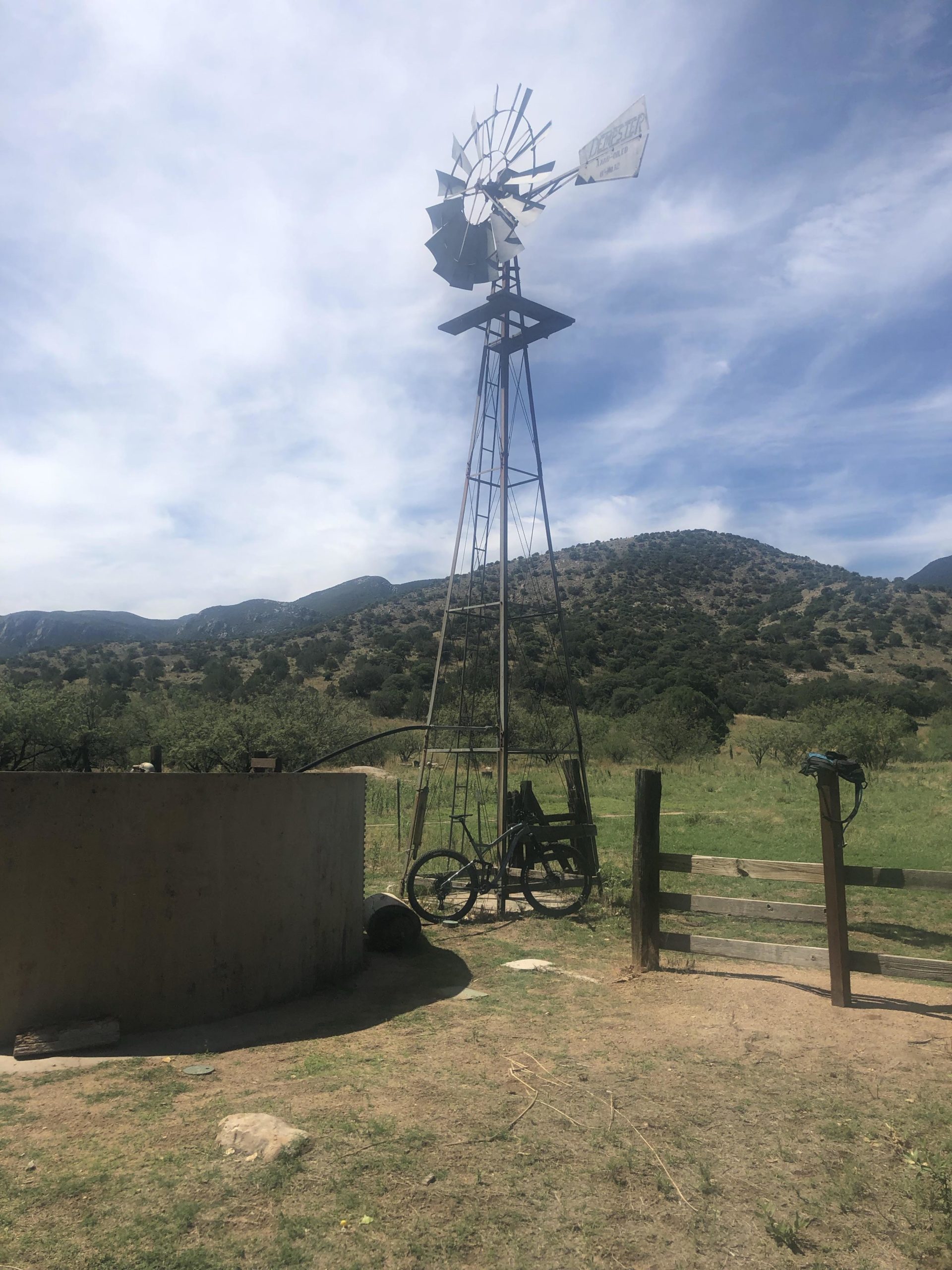 A rustic scene featuring a tall windmill against a backdrop of rolling hills and a blue sky with scattered clouds. In the foreground, there is a circular, concrete water trough, a bicycle leaning against a metal structure, and a wooden fence gate. The landscape is dotted with green grass and sparse trees, creating a serene, rural atmosphere. Brown Canyon mountain bike trail.