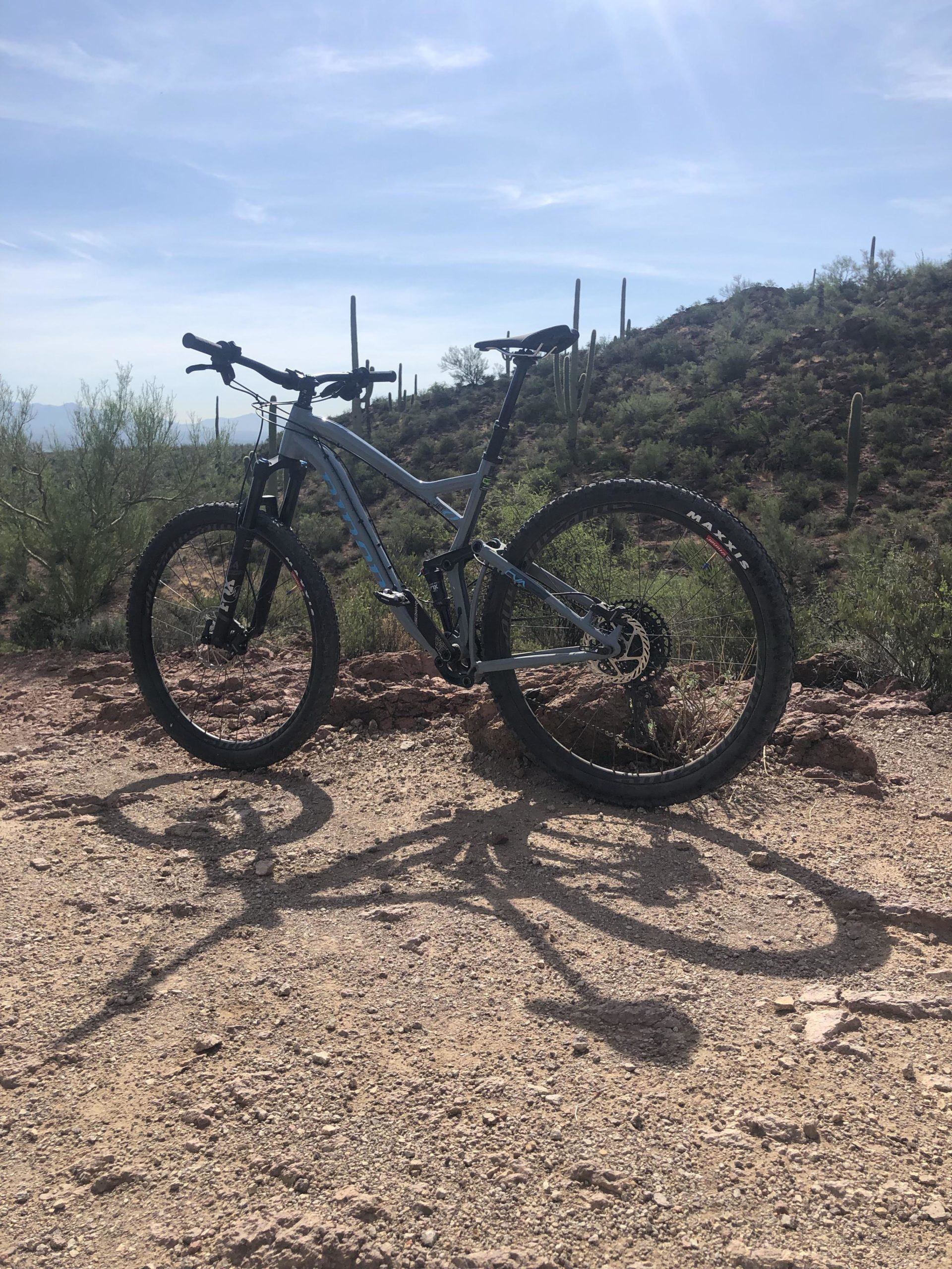 A mountain bike parked on a rocky trail with a scenic view of distant hills and cacti in the background, under a bright blue sky. The bike casts a long shadow on the gravel surface. Sweetwater Preserve mountain bike trail.