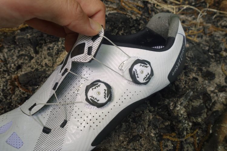 Close-up of a white cycling shoe being adjusted, featuring a unique dial closure system. The shoe displays a sleek design with textured patterns and ventilation holes, resting on a rocky surface. A hand is holding one of the dials, indicating preparation for use.
