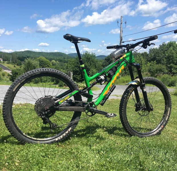 Kona Process 134: A vibrant green mountain bike parked on grassy terrain, with a scenic backdrop of rolling hills and a blue sky dotted with clouds. The bike features thick tires, a suspension system, and is positioned at an angle to highlight its design.