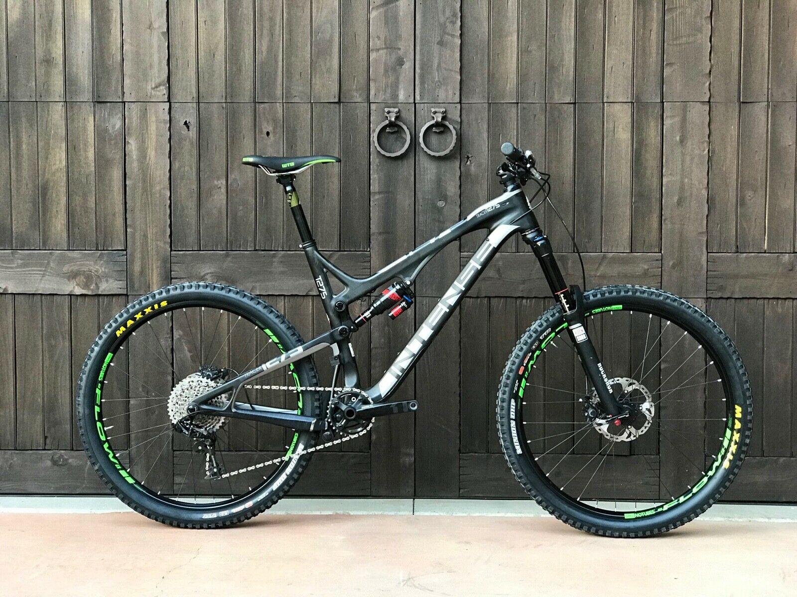 Intense Tracer 275c: Mountain bike leaning against a wooden wall, featuring a black frame with silver accents, green details on the tires and seat, and a modern design suitable for off-road riding. The background consists of large wooden doors with metal handles.