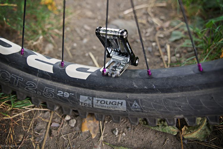 A close-up view of a mountain bike tire with a focus on the rim and spokes. The tire features the branding "TCS TOUGH HIGH GRIP" and a size label of "2.5 29". A metal bike part is attached to the spokes, showcasing its design and attachment mechanism. The surrounding area includes dirt and fallen leaves, hinting at an outdoor setting.