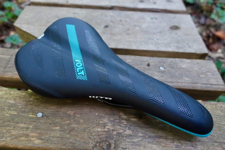 A black bicycle saddle with a blue and white "Volt" logo, featuring textured strips for enhanced grip, resting on a wooden surface in a natural outdoor setting.