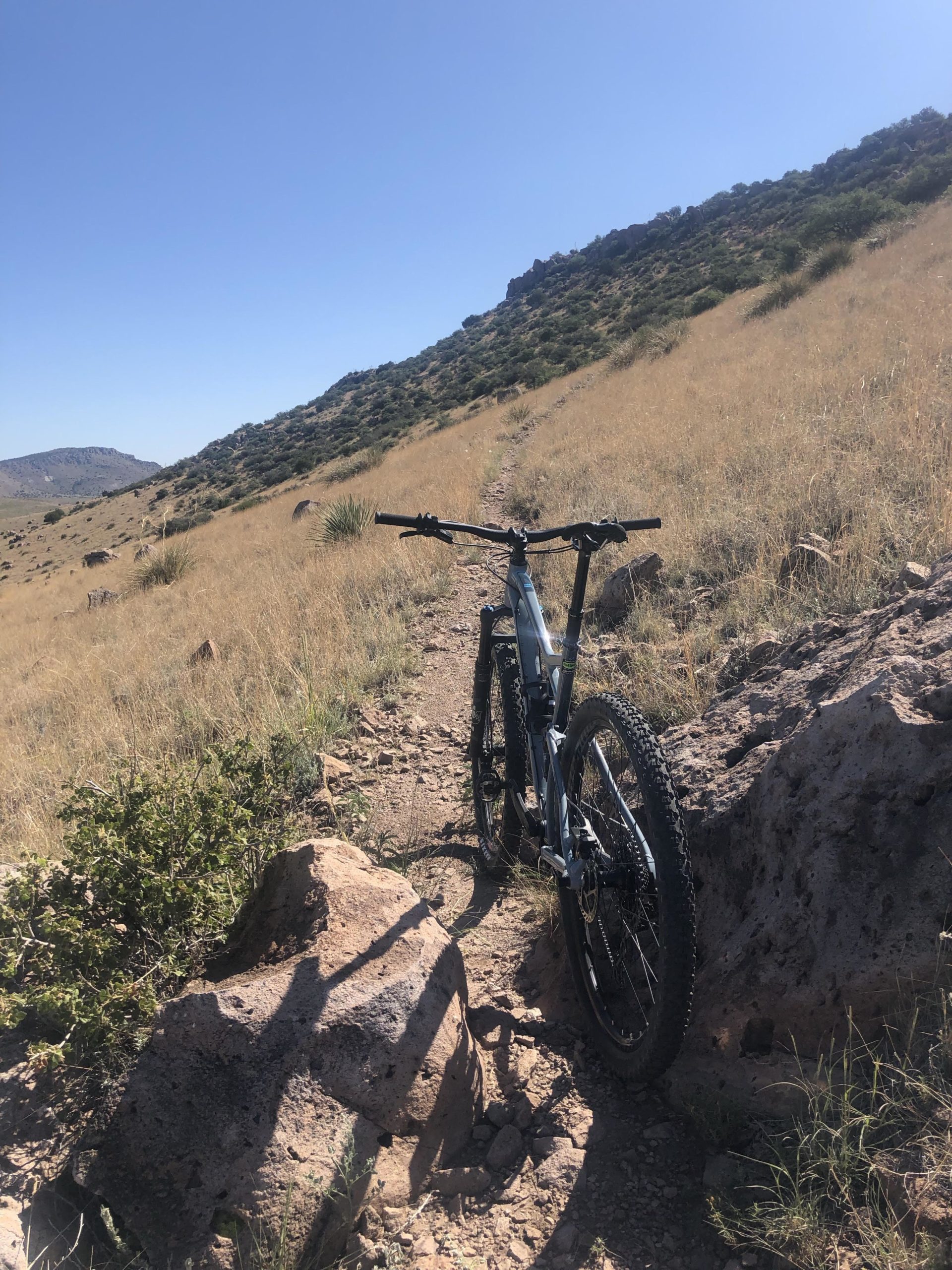 A mountain bike leaning against a large rock on a dirt trail, surrounded by tall grasses and shrubs, with a hilly landscape in the background under a clear blue sky. City Of Rocks mountain bike trail.
