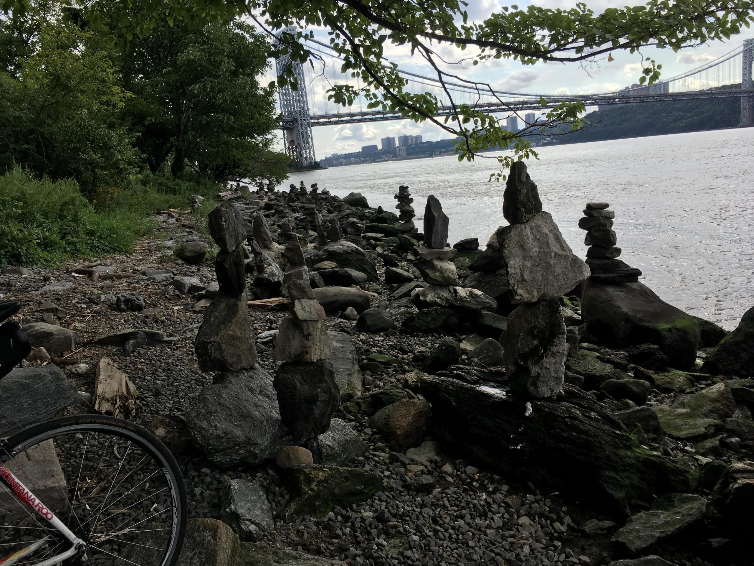 A scenic riverside view featuring a series of stacked stones along a rocky shoreline, with green foliage in the background and a bridge spanning across the water in the distance. A bicycle is partially visible in the foreground. The sky is partly cloudy. West Street Greenway mountain bike trail.
