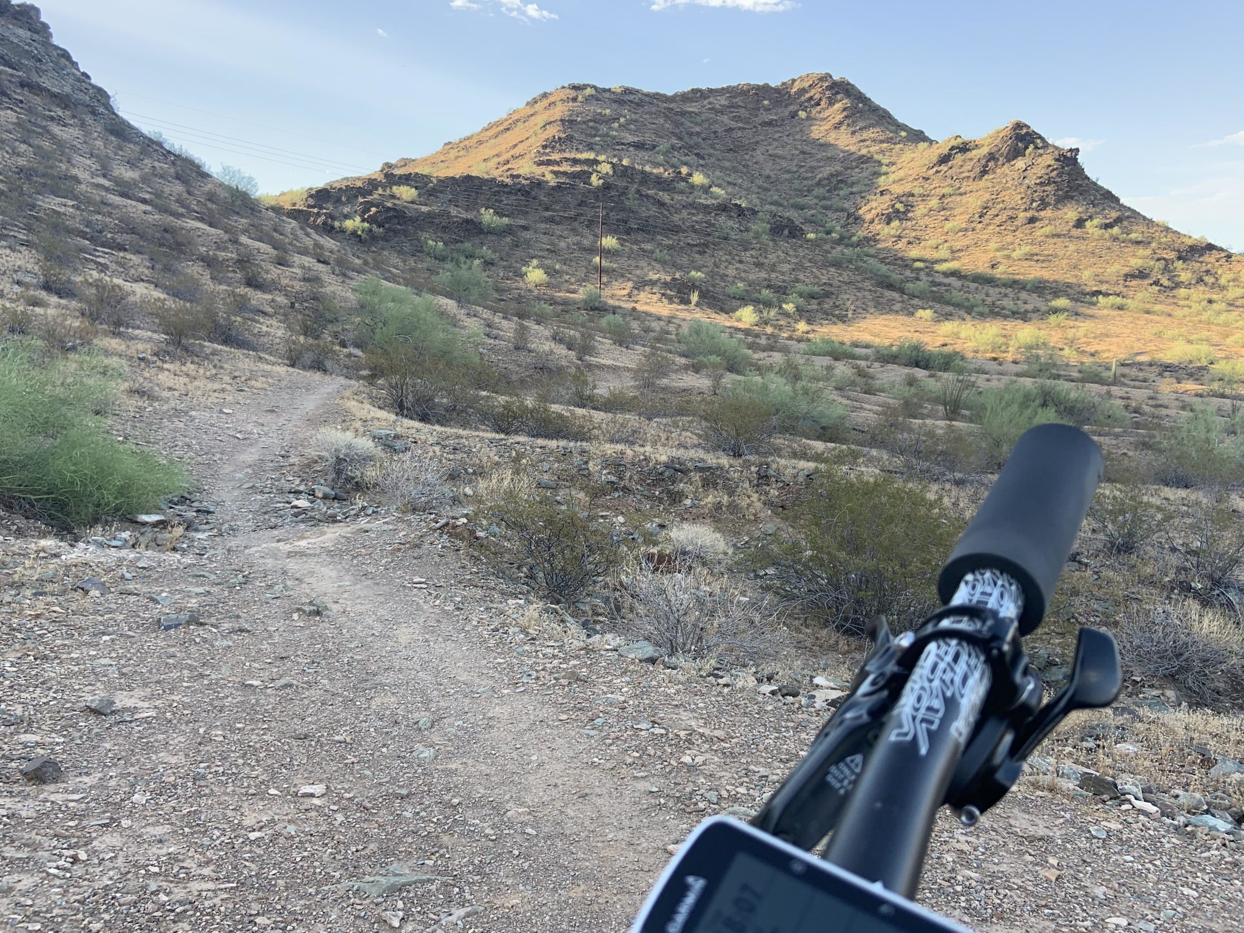 A view of a rocky dirt trail winding through a desert landscape, with low shrubs and hills in the background. The image includes the handlebars and display of a mountain bike in the foreground, suggesting an outdoor biking adventure. Trail #100 mountain bike trail.