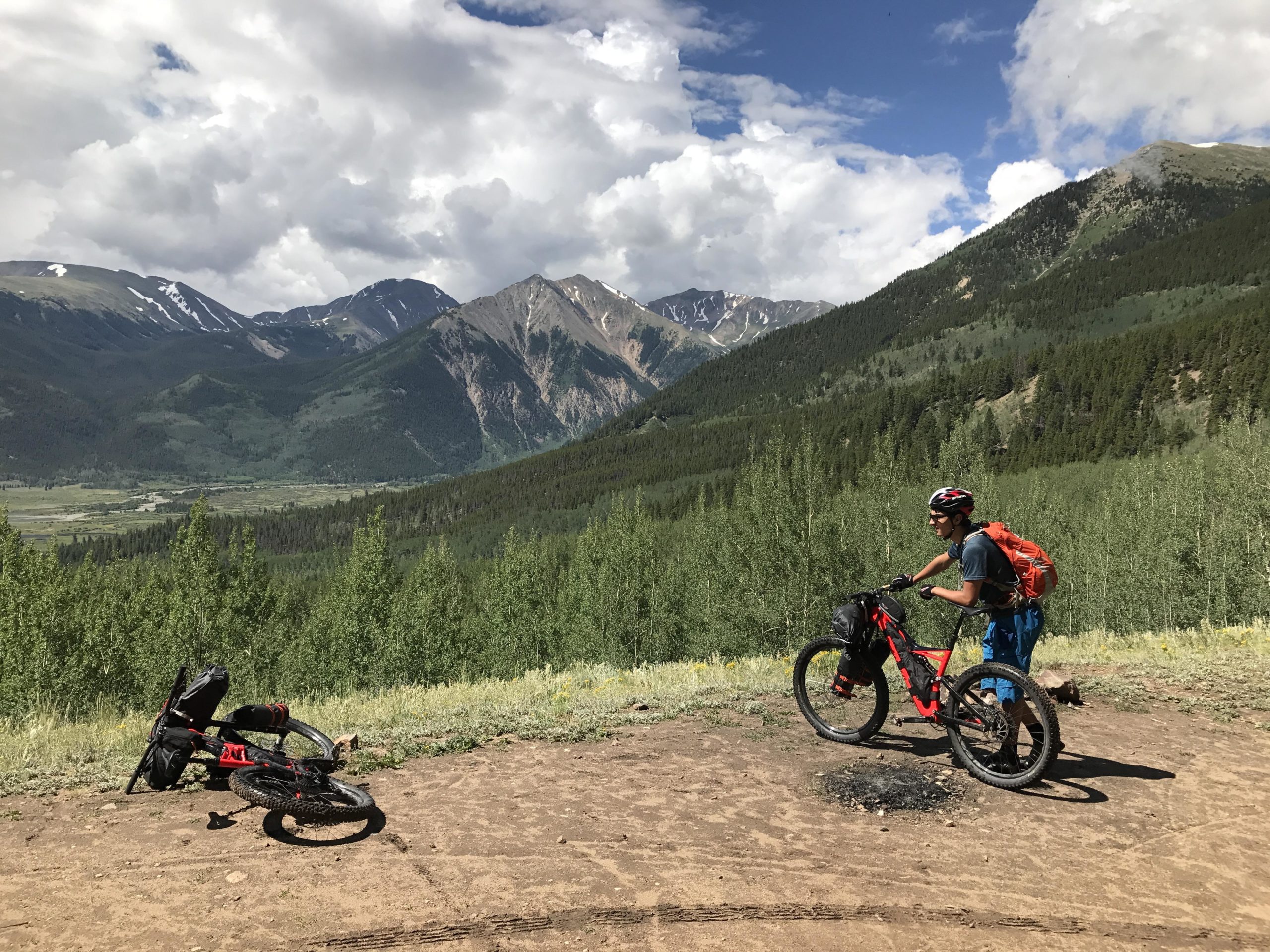 A person in outdoor gear stands next to a mountain bike on a dirt trail, surrounded by lush green trees and mountains in the background. The sky is partly cloudy with patches of blue. A bike is lying on the ground nearby, indicating a stop in a scenic mountainous area. Colorado Trail mountain bike trail.