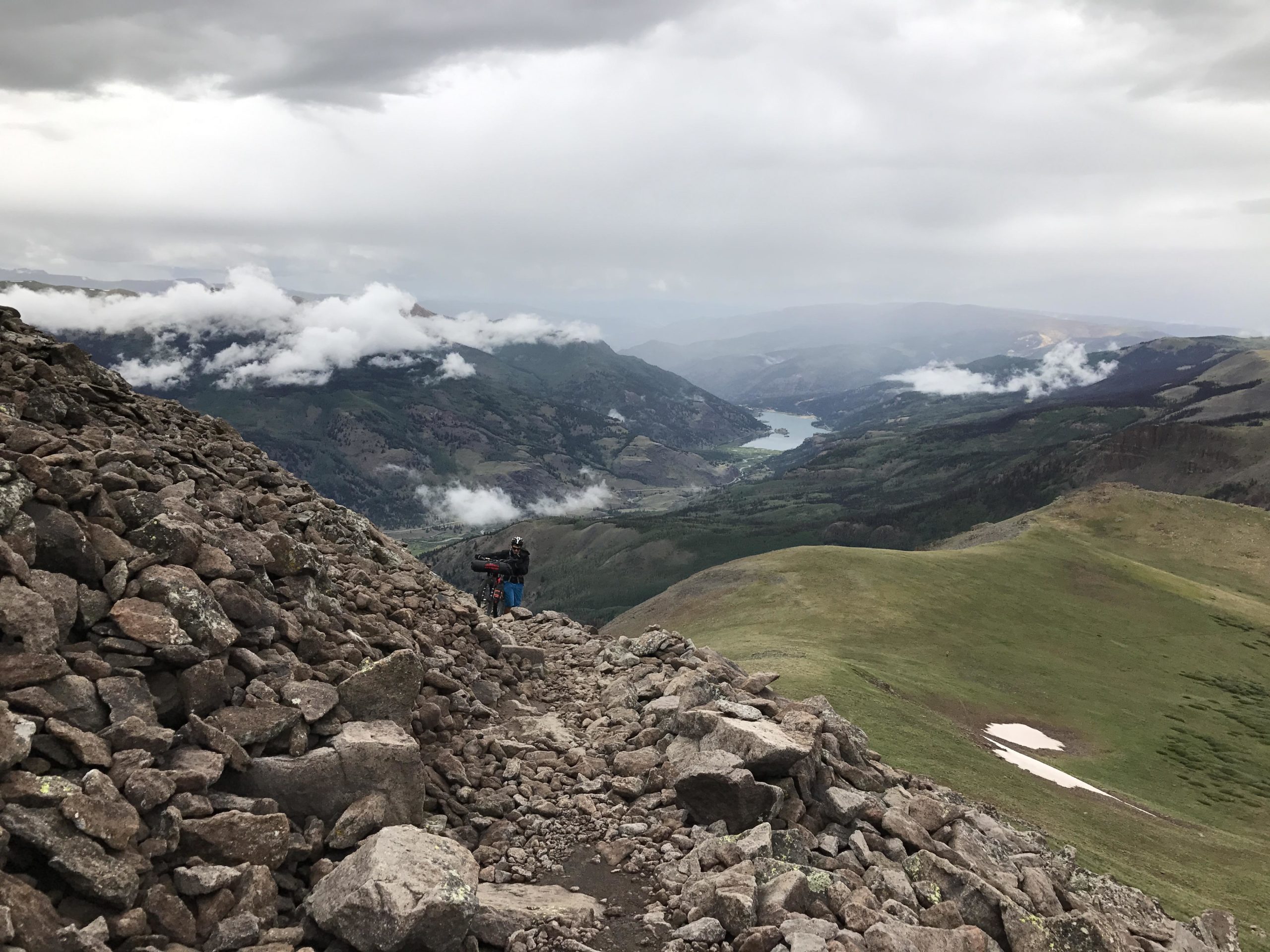 A rugged mountain landscape featuring a rocky trail leading up to an elevated viewpoint. In the foreground, a hillside of loose stones is visible, while in the background, a person stands with a bike, gazing out at a valley filled with greenery and a lake, under a cloudy sky. Colorado Trail mountain bike trail.