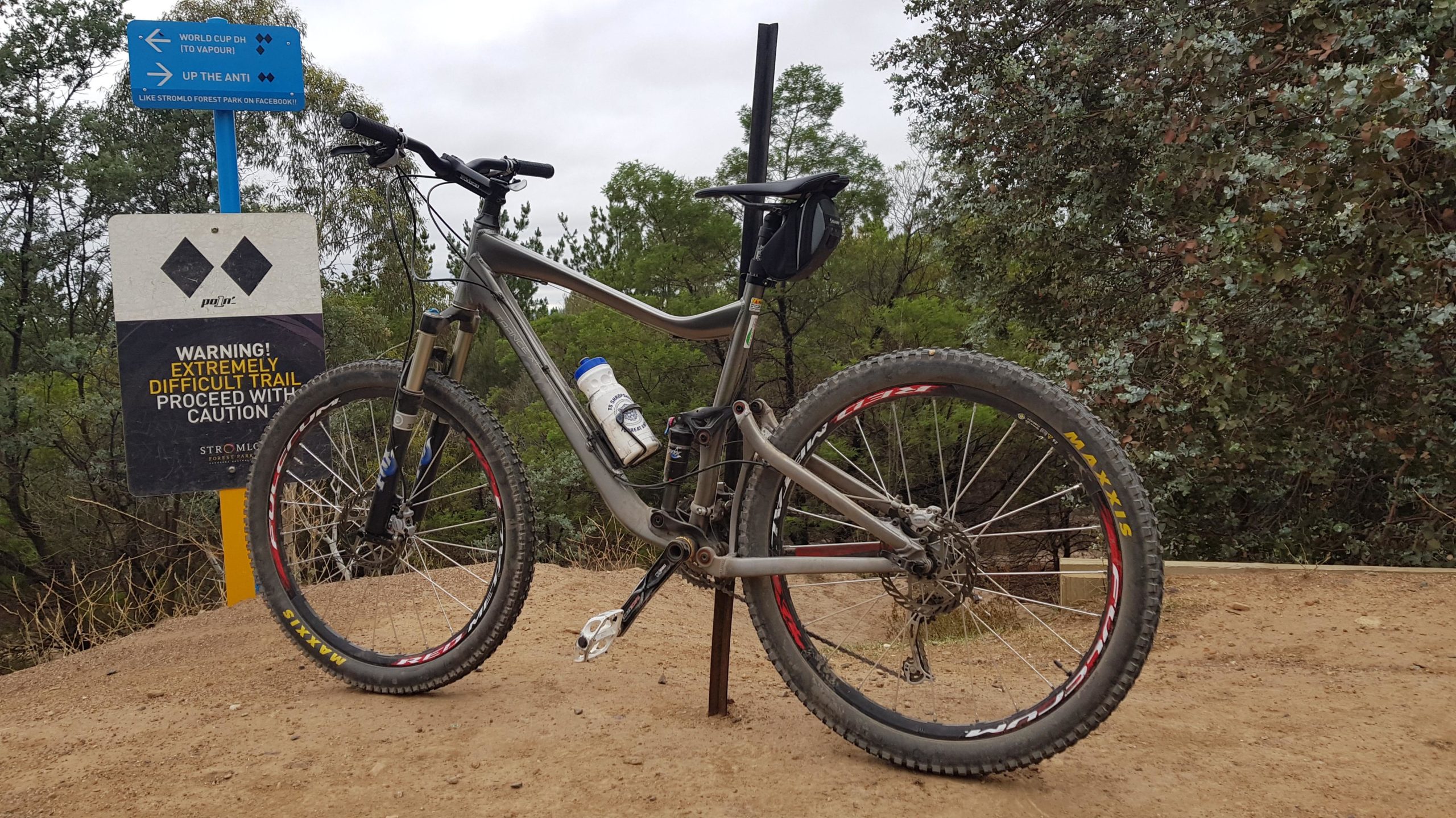 A mountain bike parked on a dirt trail, with a warning sign indicating an extremely difficult trail ahead. The bike features thick tires and a water bottle attached to its frame. In the background, trees and shrubs are visible under a cloudy sky. Stromlo Forest Park mountain bike trail.