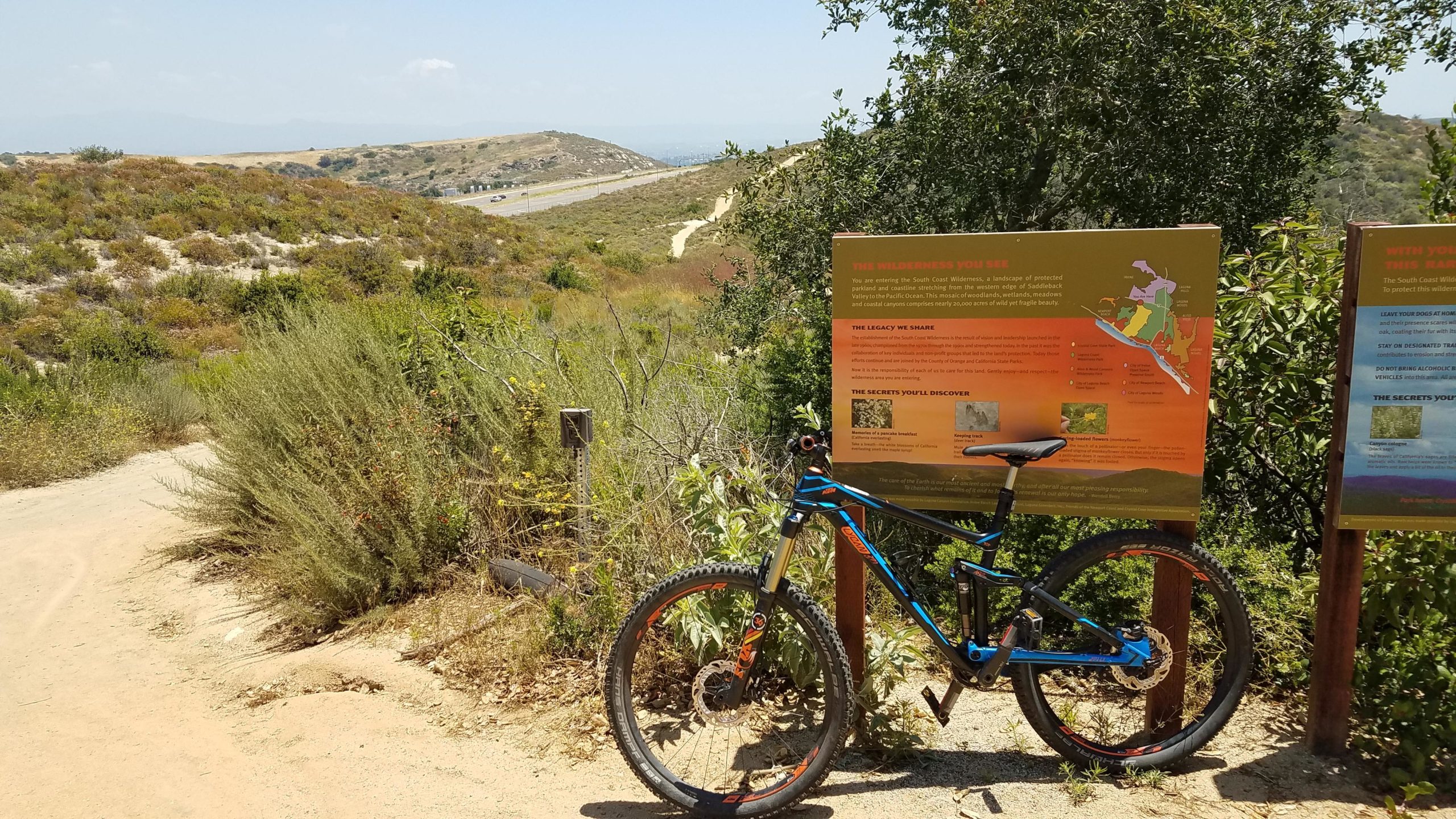 A mountain bike resting on a sandy trail surrounded by lush greenery, with informational signs about the South Coast Wilderness in the background. The landscape features rolling hills under a clear blue sky. Laguna Coast Wilderness Park mountain bike trail.