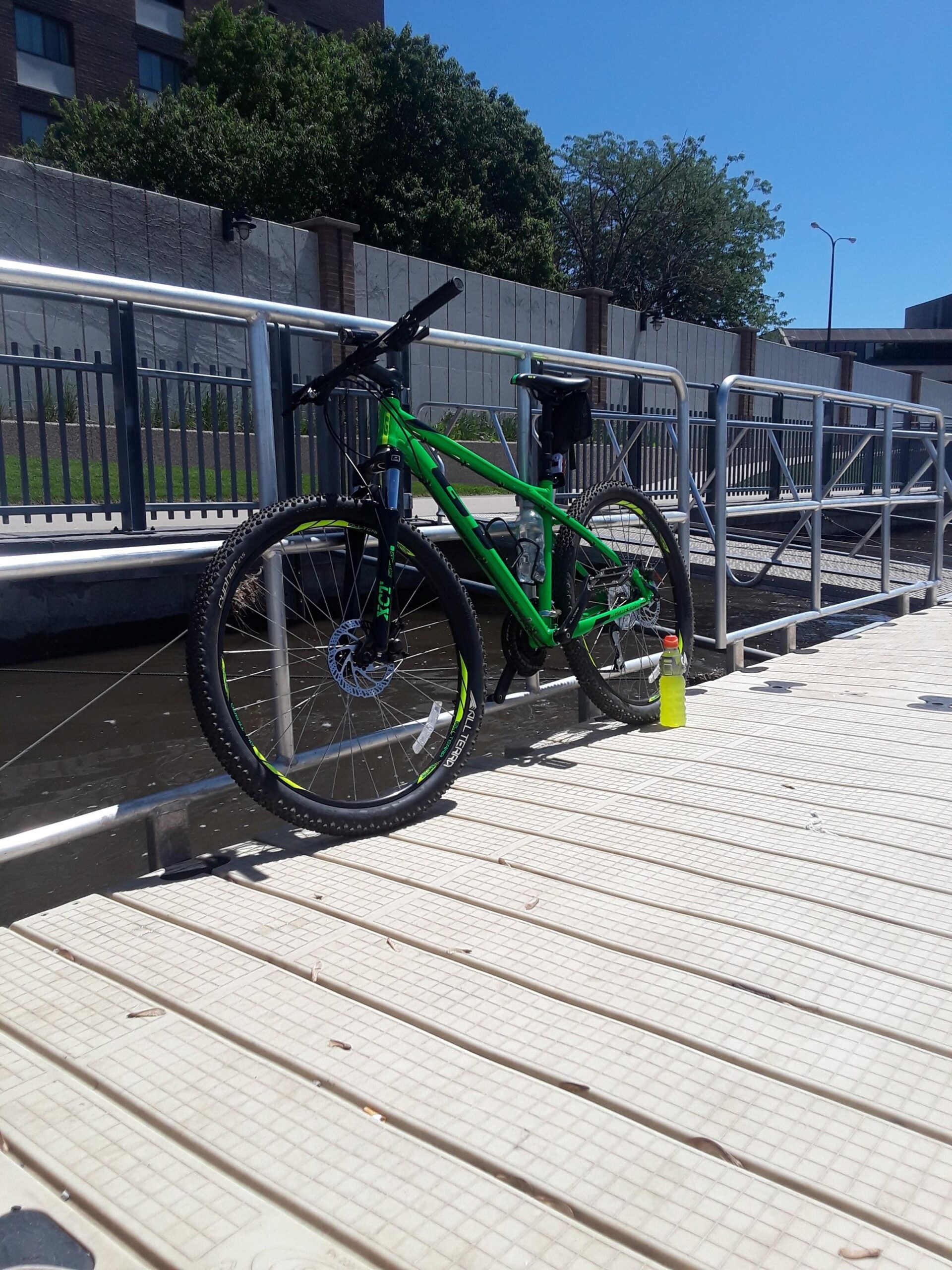 GT Aggressor Pro: A green mountain bike is parked on a wooden dock beside a body of water, with a clear blue sky overhead. A water bottle is placed next to the bike. In the background, a building and trees are visible.