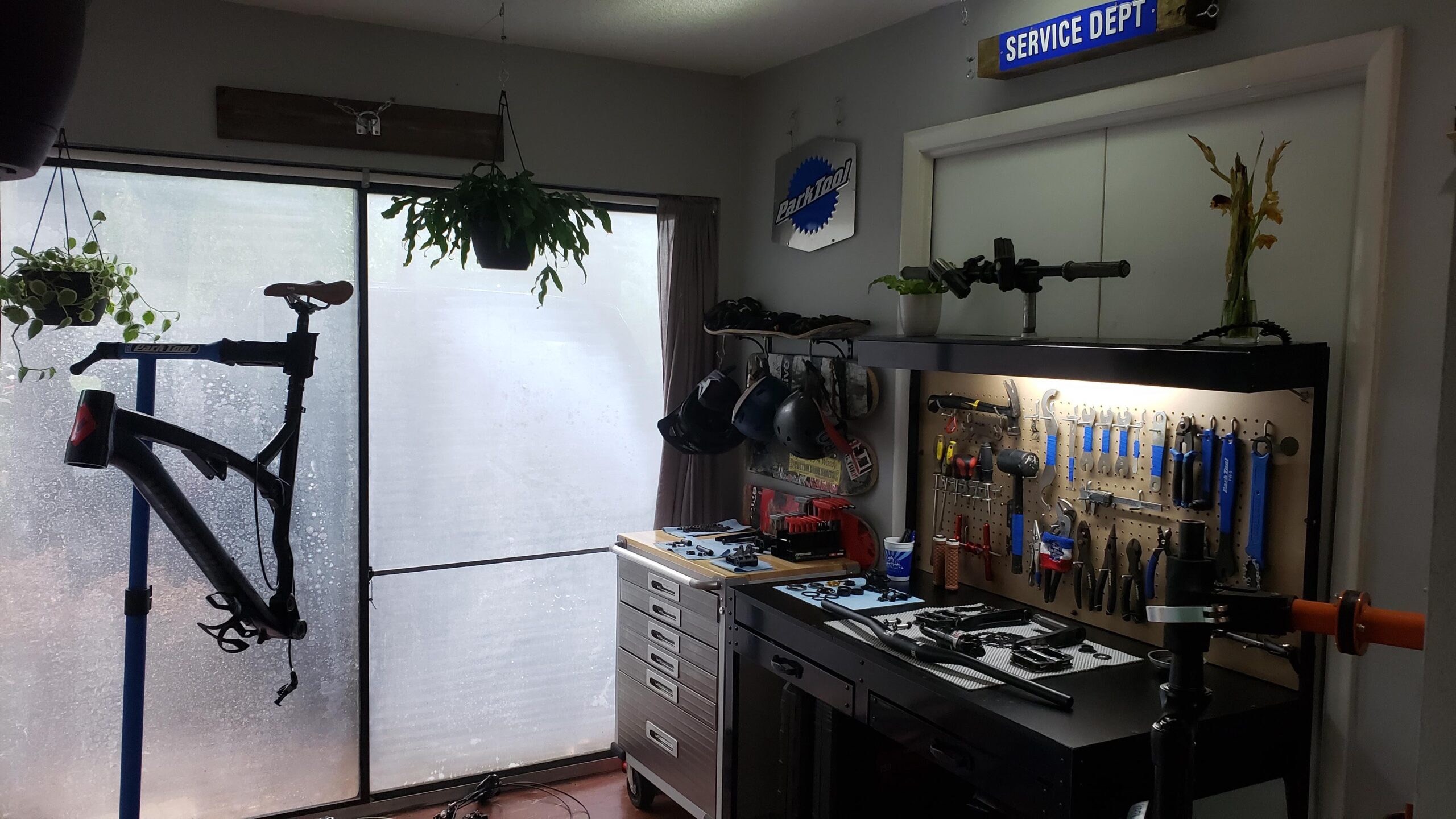 Diamondback Diamondback Release 1 27.5 2017: A well-organized bike repair workshop featuring a bike stand with a black bike frame, a pegboard adorned with various hand tools in shades of blue, and a tool chest. The room has large windows partially covered by curtains, with hanging plants above and a "Service Department" sign on the door. A small vase with flowers adds a decorative touch to the workspace.