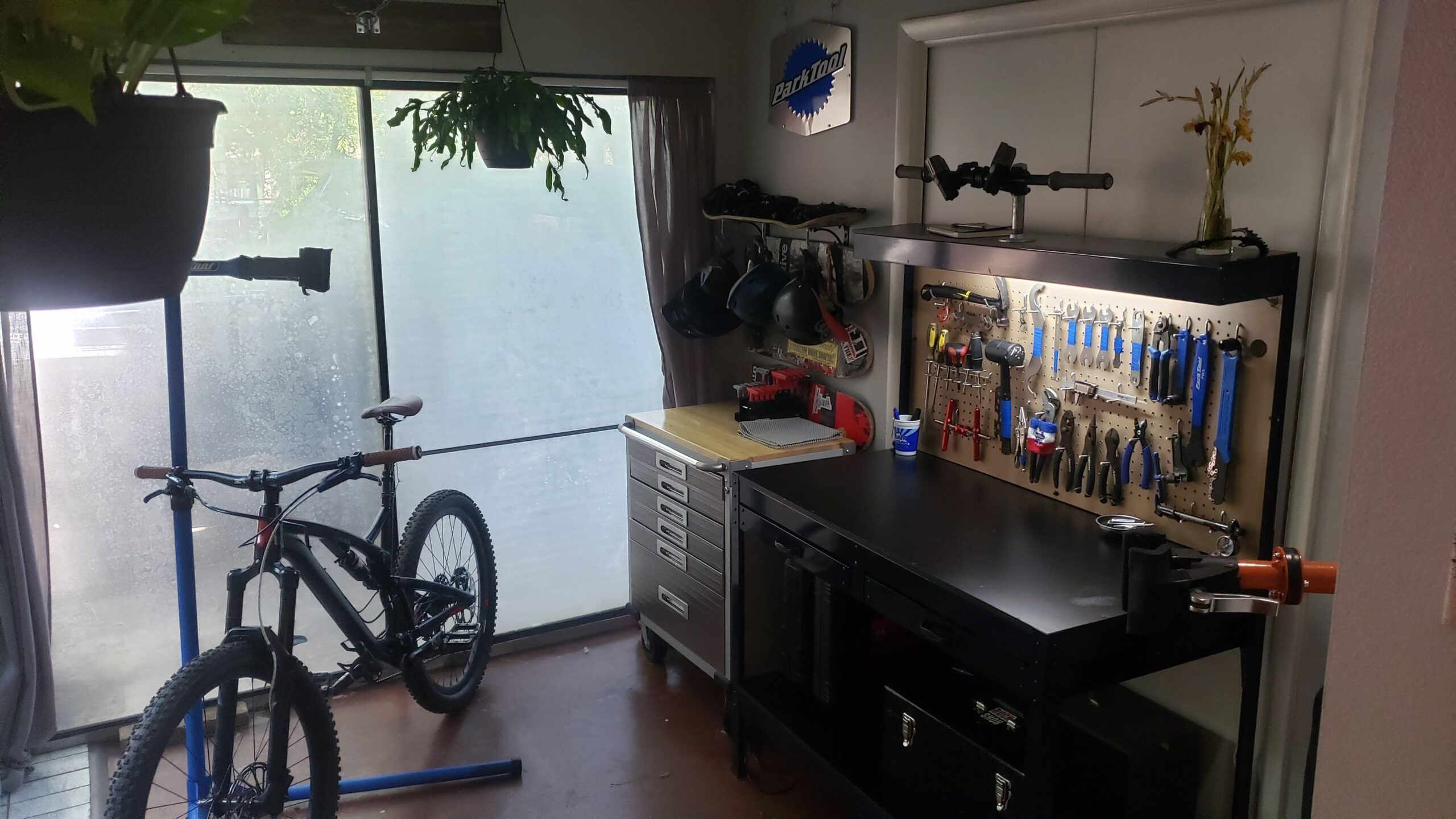Diamondback Diamondback Release 1 27.5 2017: A well-organized bike maintenance area featuring a mountain bike on a repair stand, a workbench with various tools neatly hung on a pegboard, and a storage cart with drawers. Large windows provide natural light, and there are potted plants hanging near the window.