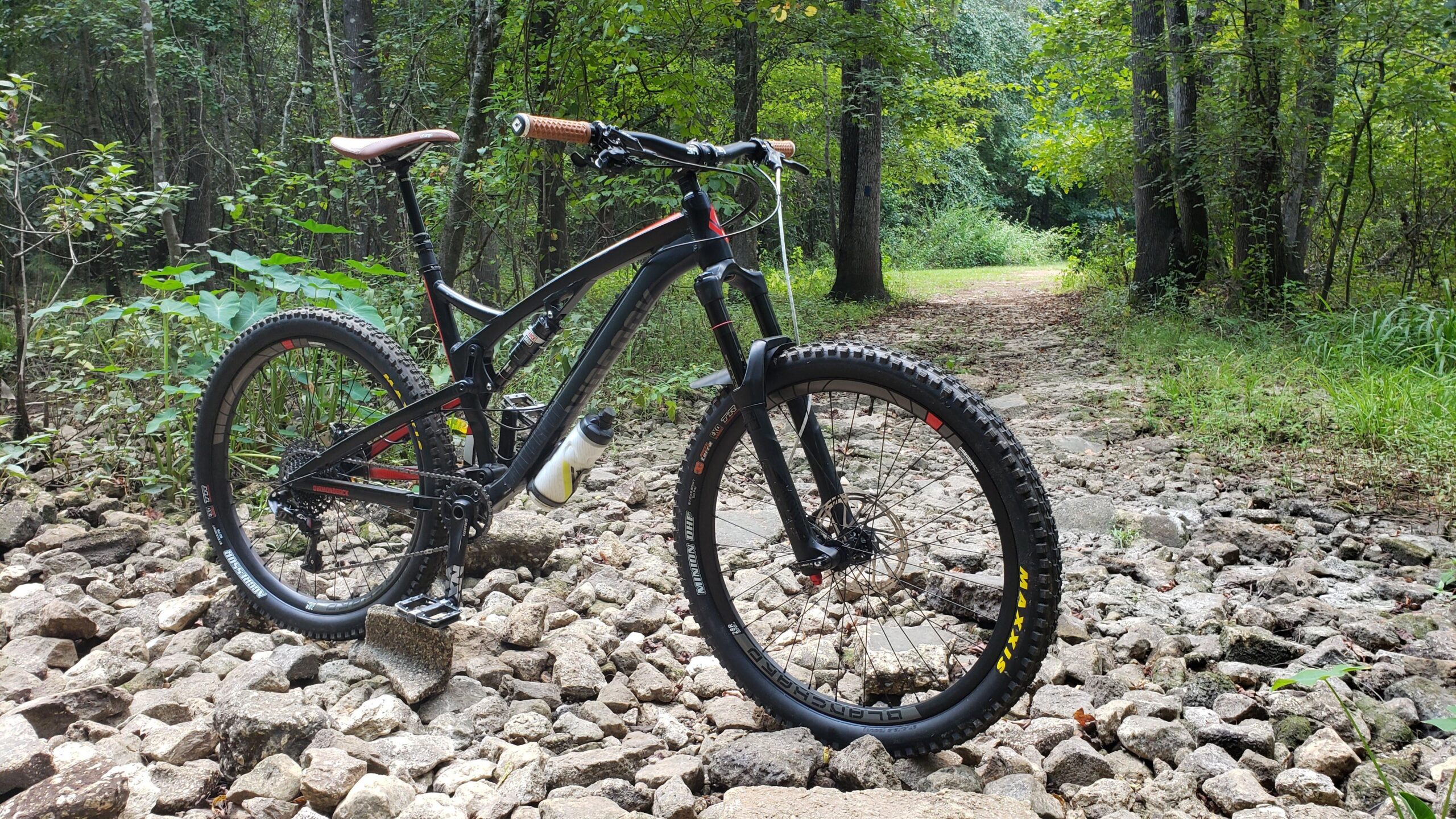 Diamondback Diamondback Release 1 27.5 2017: A mountain bike resting on a rocky trail surrounded by lush green foliage and trees, with a clear path visible in the background. The bike features a sleek black frame with brown handlebars and a water bottle attached.