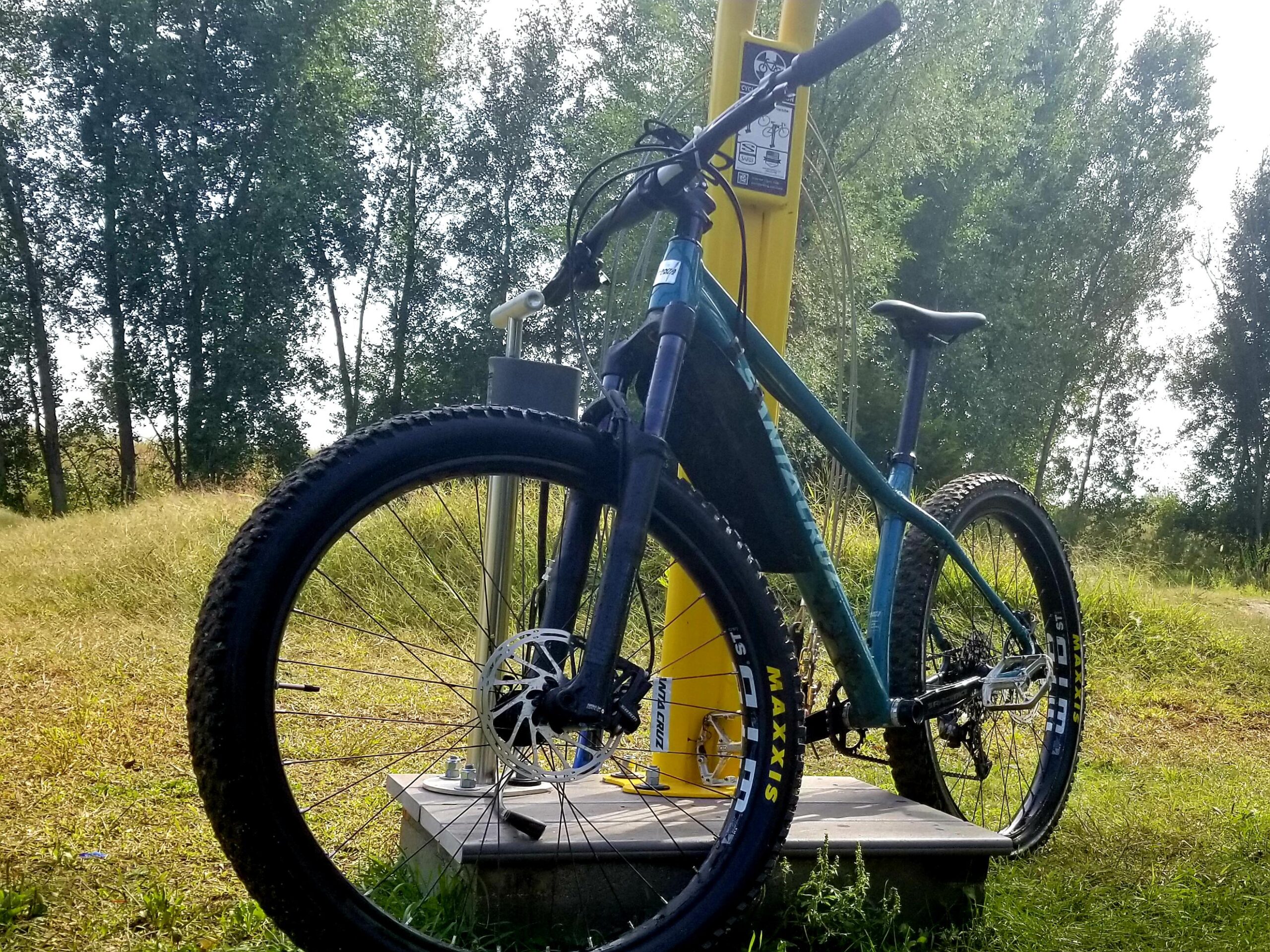 Santa Cruz Chameleon: A mountain bike parked beside a yellow bike maintenance station, set against a backdrop of green trees and grassy terrain. The bike features thick tires and a sturdy frame, indicating it's built for rugged trails.