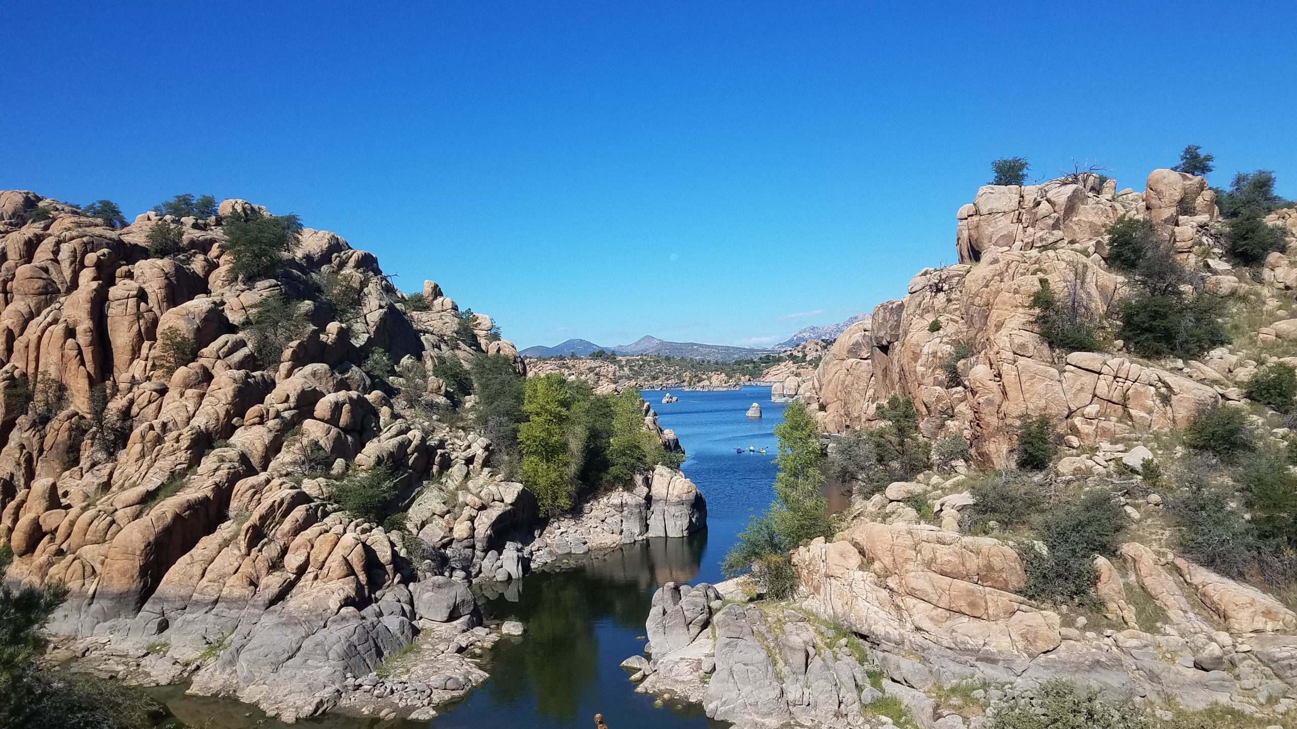 A scenic view of rocky terrain with trees alongside a tranquil blue lake, under a clear blue sky. The landscape features large, rugged rock formations along the water