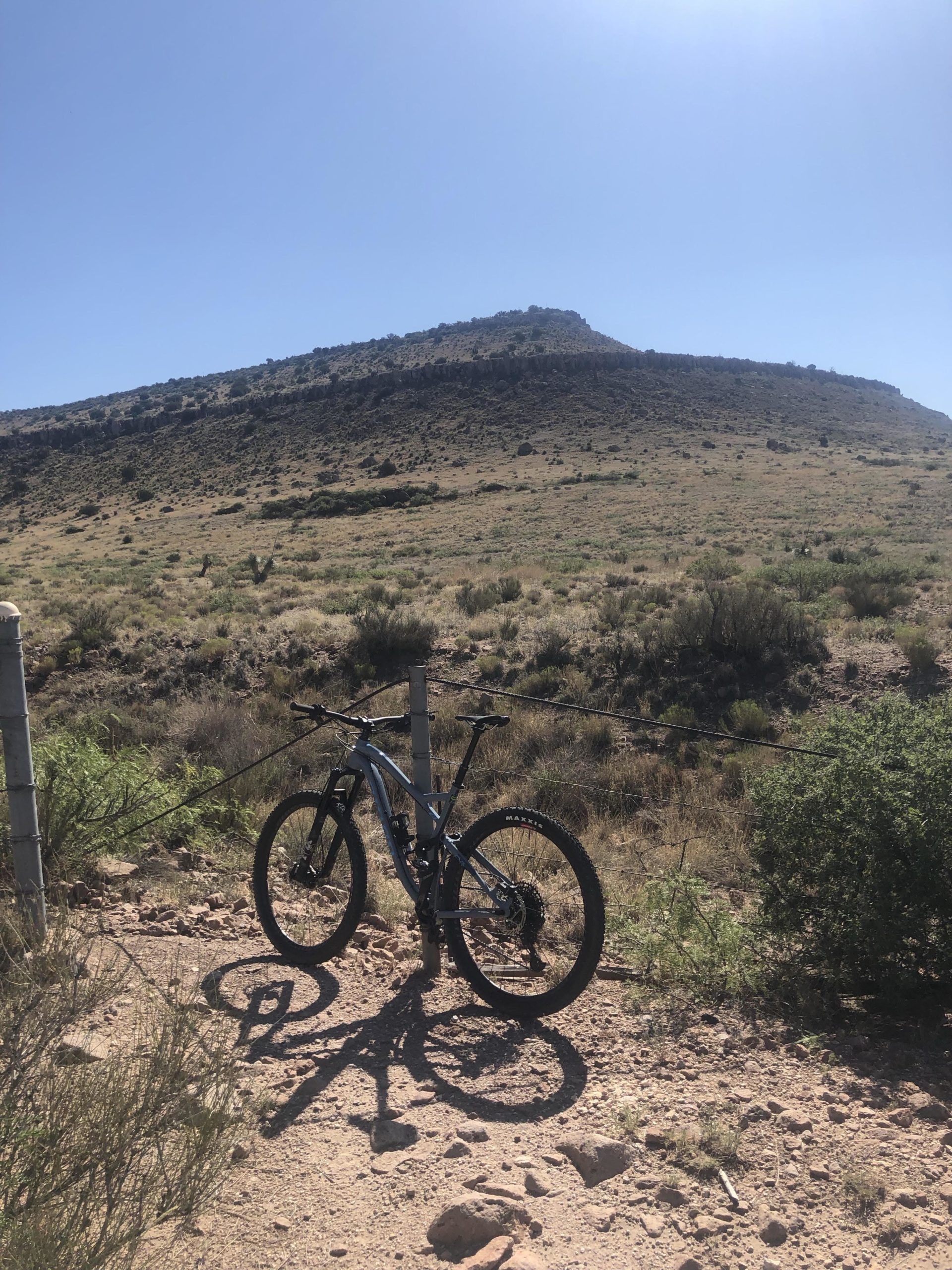 A mountain bike parked by a dirt trail, with a hilly landscape in the background under a clear blue sky. The foreground features sparse vegetation and rocky terrain. City Of Rocks mountain bike trail.