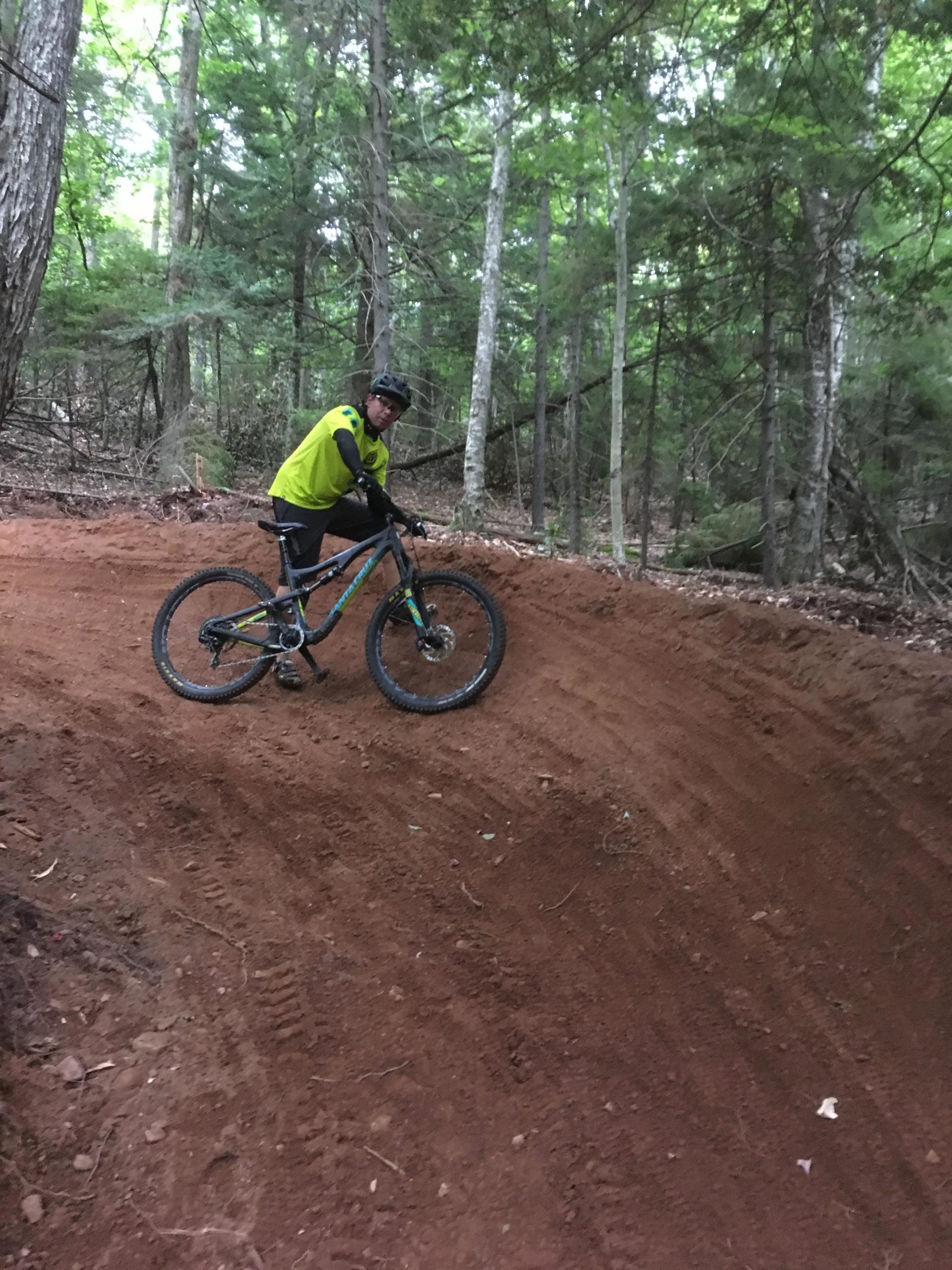 A person wearing a bright yellow shirt and black pants stands next to a mountain bike on a dirt trail in a forest. They are positioned on a slope with tall trees and greenery surrounding them. Tannersville Bike Park [Hathaway Trails] mountain bike trail.