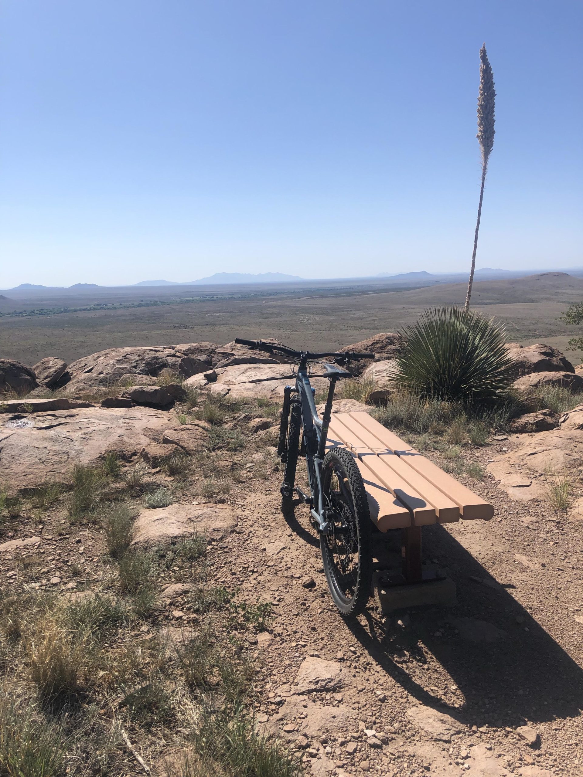 A mountain bike is parked next to a wooden bench on a rocky outcrop, overlooking a vast, open landscape with distant mountains under a clear blue sky. Sunlight casts shadows on the ground, highlighting the natural surroundings of grass and rocks. City Of Rocks mountain bike trail.