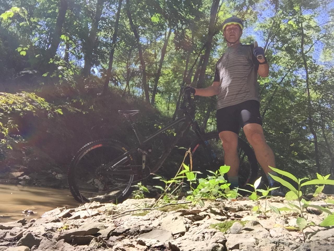 A person in athletic clothing stands beside a mountain bike next to a creek, set in a lush green forest. Sunlight filters through the trees, illuminating the scene. The individual is giving a thumbs-up gesture, indicating enjoyment of the outdoor activity. England Idlewild Mountain Biking Park mountain bike trail.