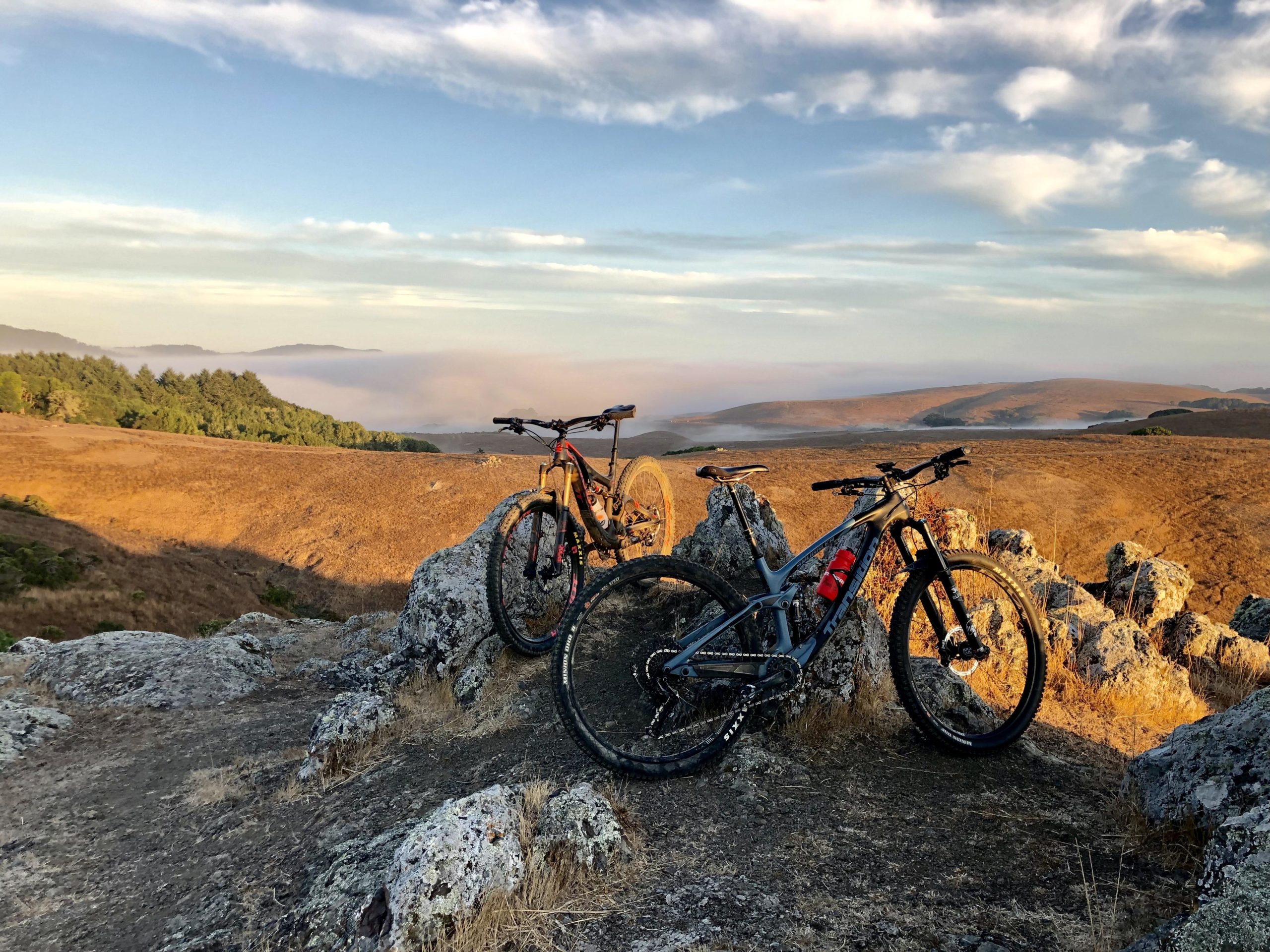 Two mountain bikes rest on a rocky outcrop, overlooking a vast, golden landscape with hills and a misty valley in the background. The sky is painted with soft wispy clouds, suggesting either dawn or dusk. The scene conveys a sense of adventure and tranquility in nature. Bolinas Ridge mountain bike trail.