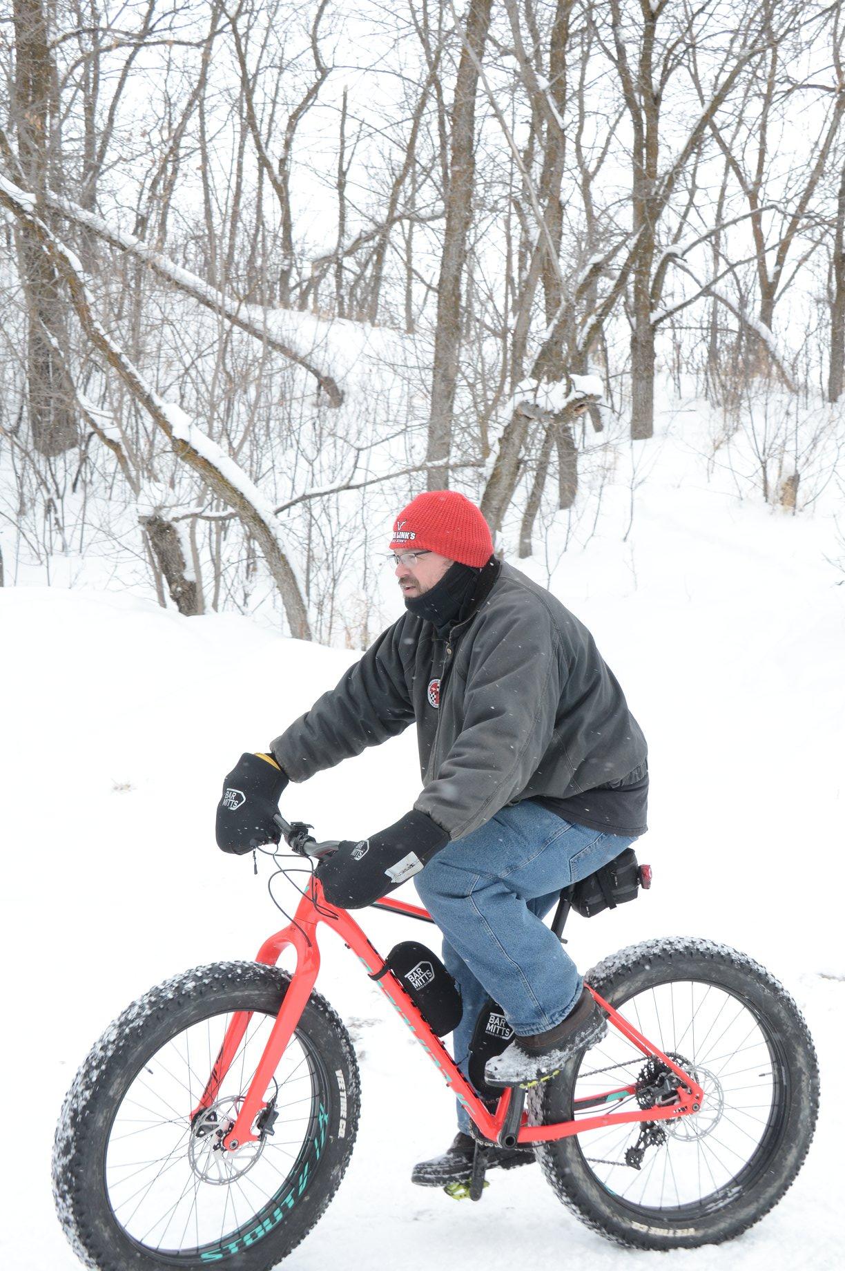 Specialized Fatboy: A man wearing a red hat, black gloves, and a dark jacket rides a fat-tire bike through a snowy landscape with bare trees in the background.