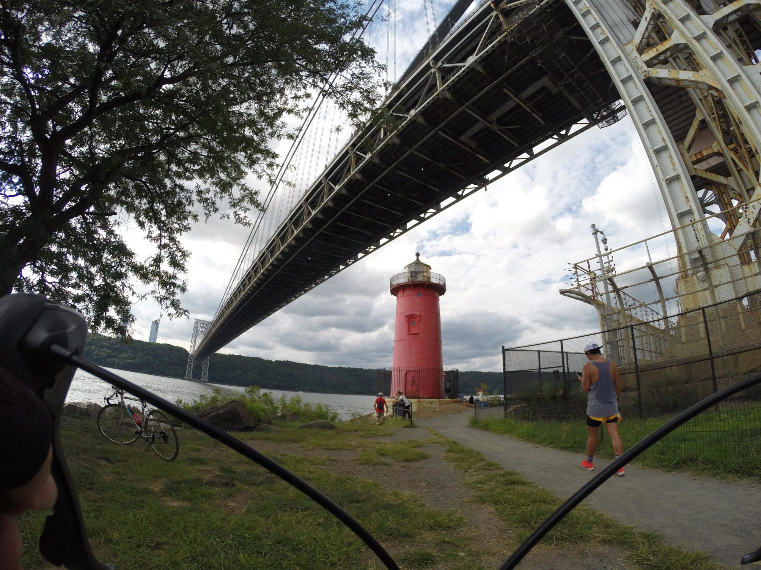 A view from the ground near the Hudson River, featuring a prominent red lighthouse under a large bridge. The scene includes a path with a person walking and a bicycle parked nearby. In the background, there are trees, a cloudy sky, and a distant building visible through the bridge's structure. West Street Greenway mountain bike trail.