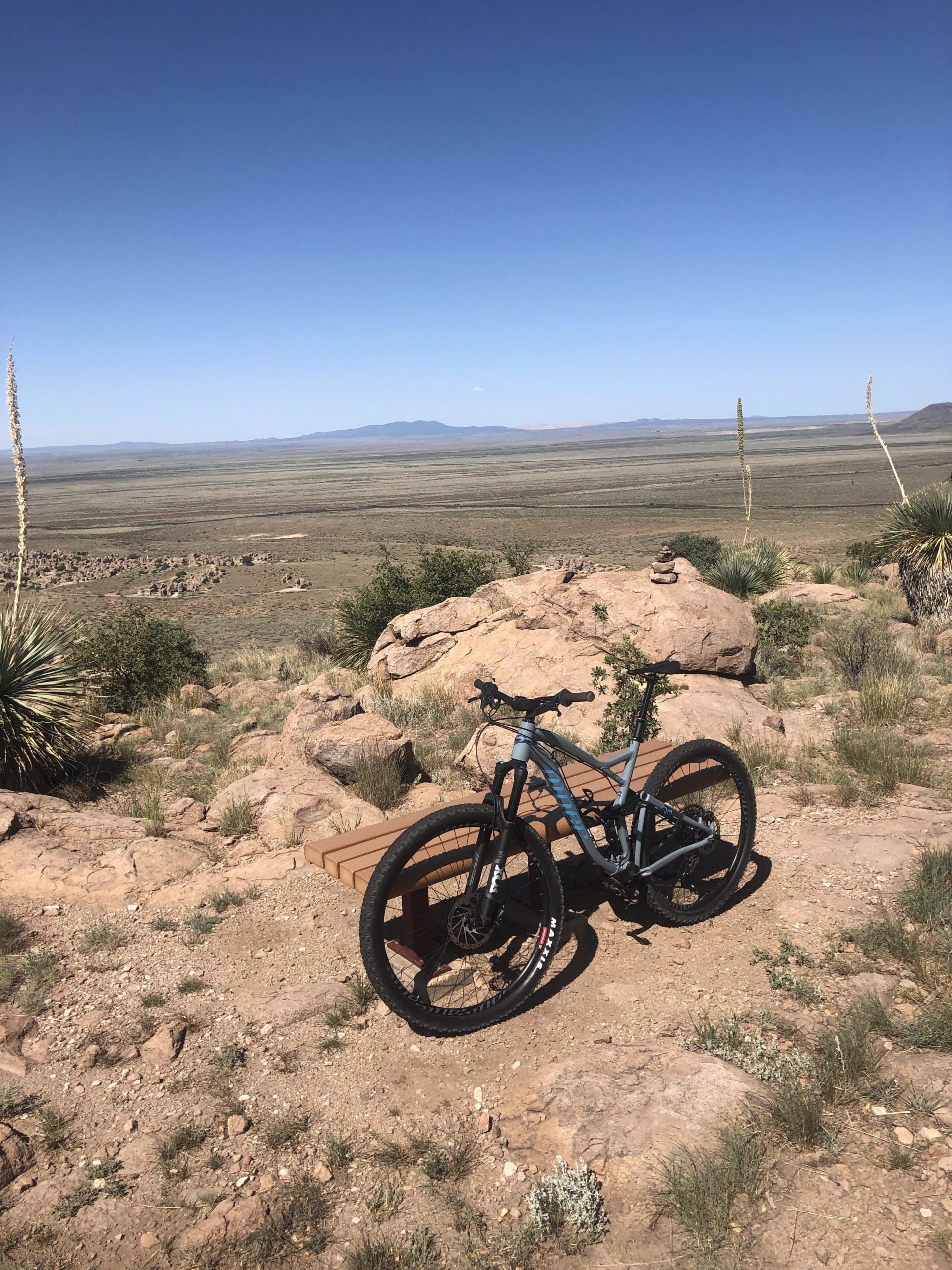 A mountain bike parked on a rocky outcrop with a vast desert landscape stretching out in the background under a clear blue sky. The scene features sparse vegetation and distant mountains, creating a serene outdoor setting. City Of Rocks mountain bike trail.