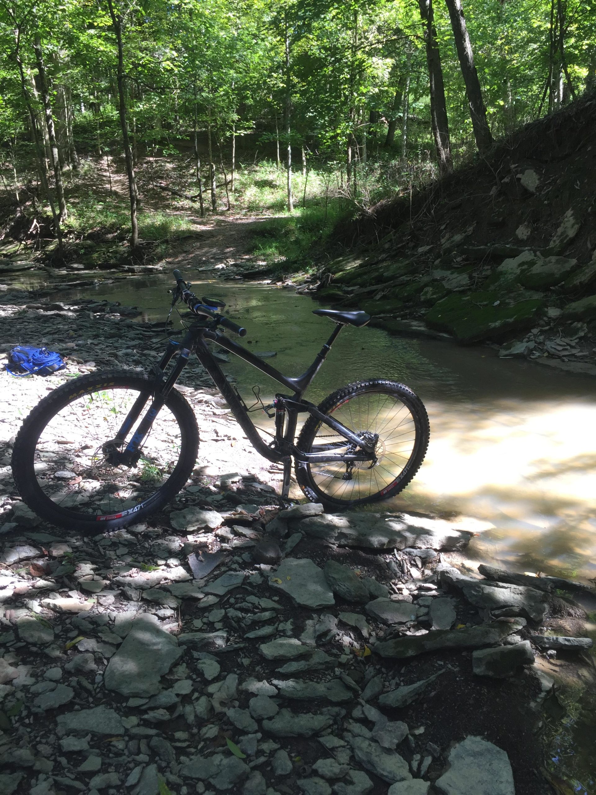 Mountain bike resting on rocky terrain beside a creek, surrounded by lush green trees and sunlight filtering through the leaves. England Idlewild Mountain Biking Park mountain bike trail.