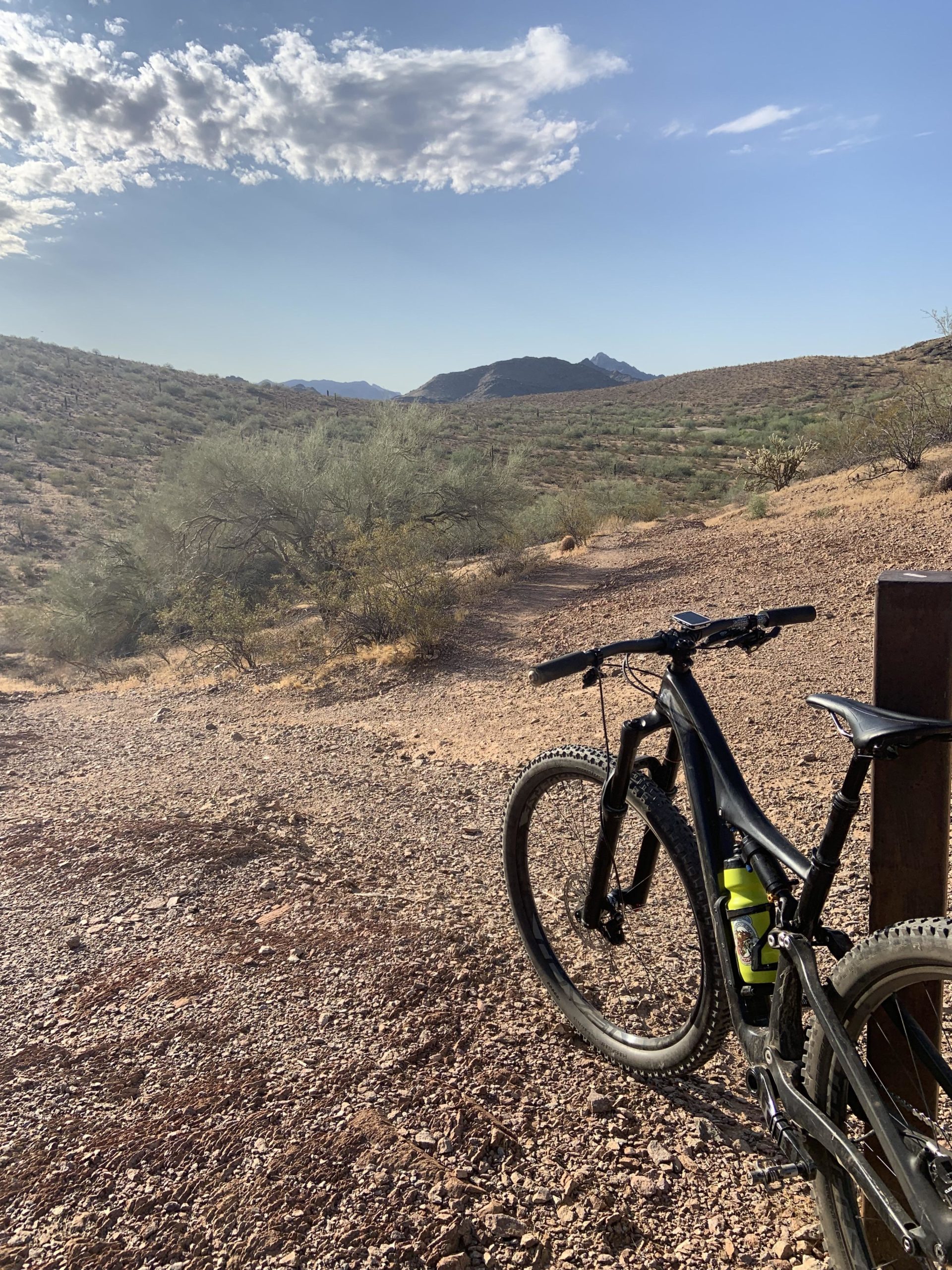 A mountain bike parked next to a trail in a desert landscape, with rolling hills and sparse vegetation under a partly cloudy sky. Mountains are visible in the background, and the terrain is rocky and dry. Trail #100 mountain bike trail.