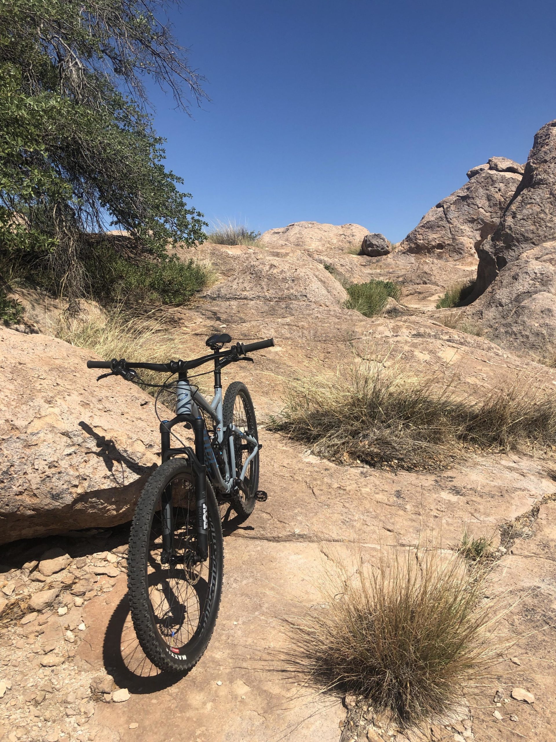 A mountain bike leaning against a rocky outcrop on a sunlit trail surrounded by sparse vegetation and a clear blue sky. City Of Rocks mountain bike trail.