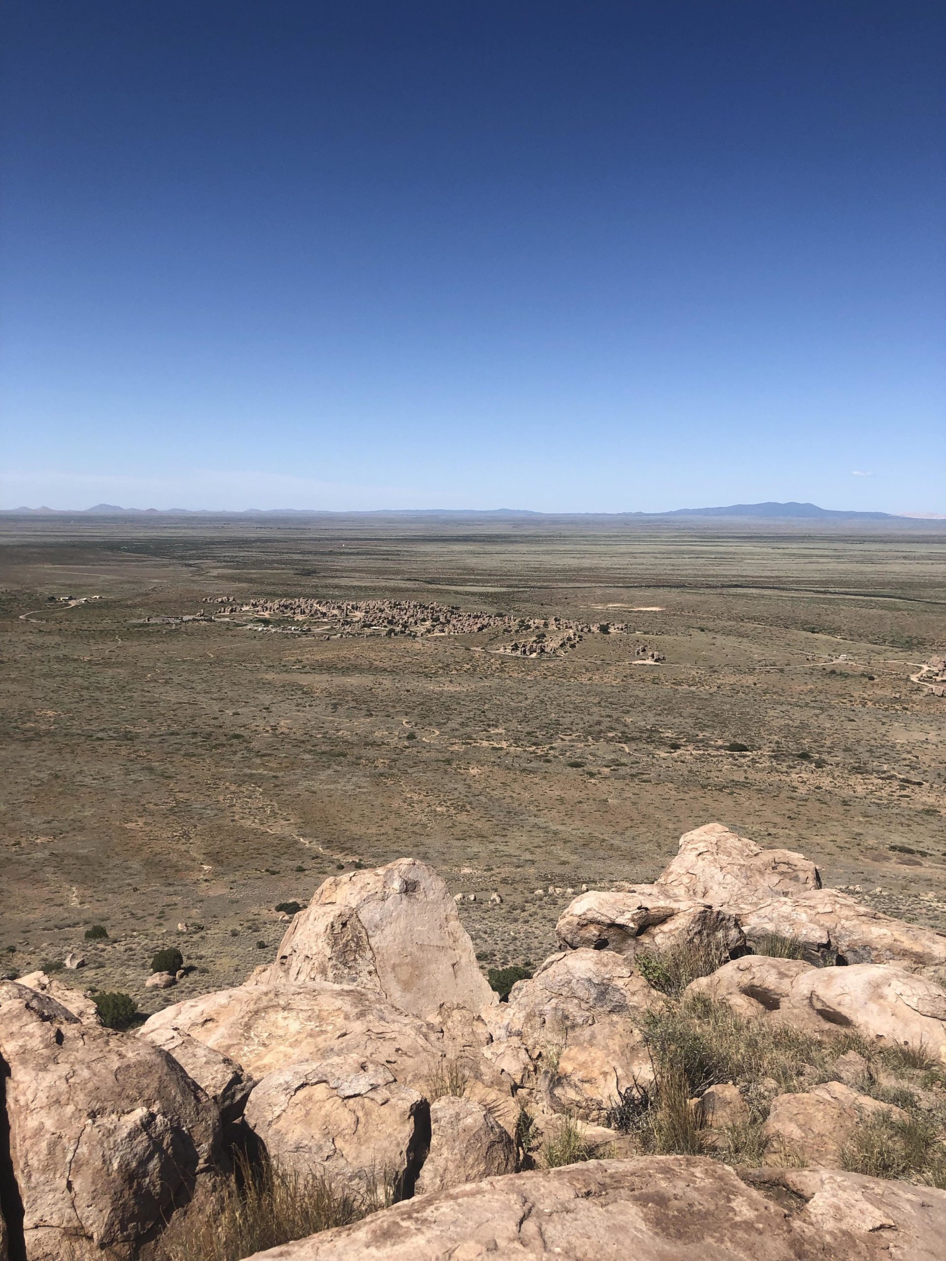 A panoramic view of a vast, arid landscape with rocky formations in the foreground and gently rolling hills in the distance under a clear blue sky. The area appears to have sparse vegetation, indicating a dry environment. City Of Rocks mountain bike trail.