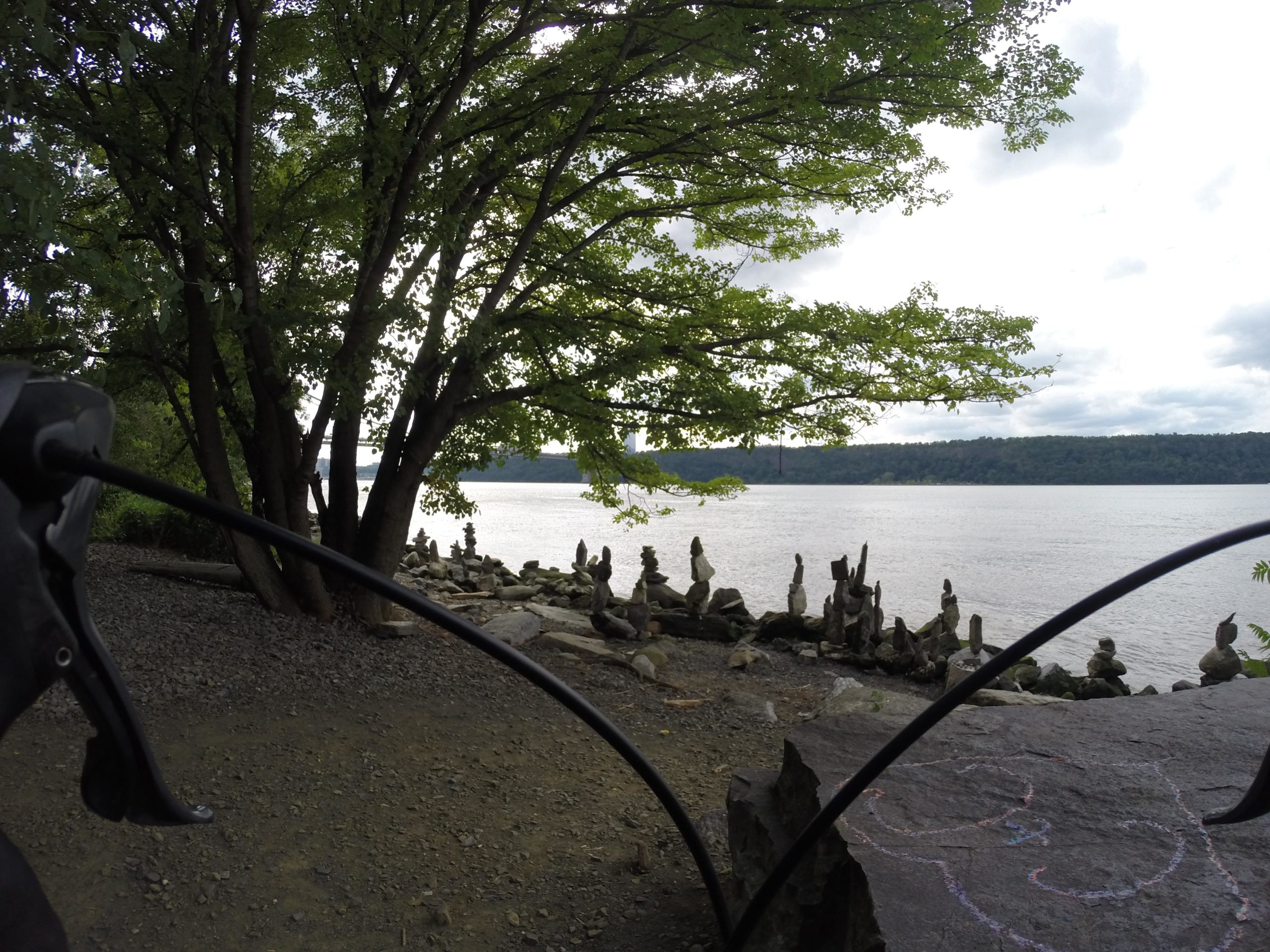 A riverside view with a tree in the foreground. The image shows a rocky shore with several carefully stacked stones along the water's edge, and a calm river in the background under a cloudy sky. West Street Greenway mountain bike trail.