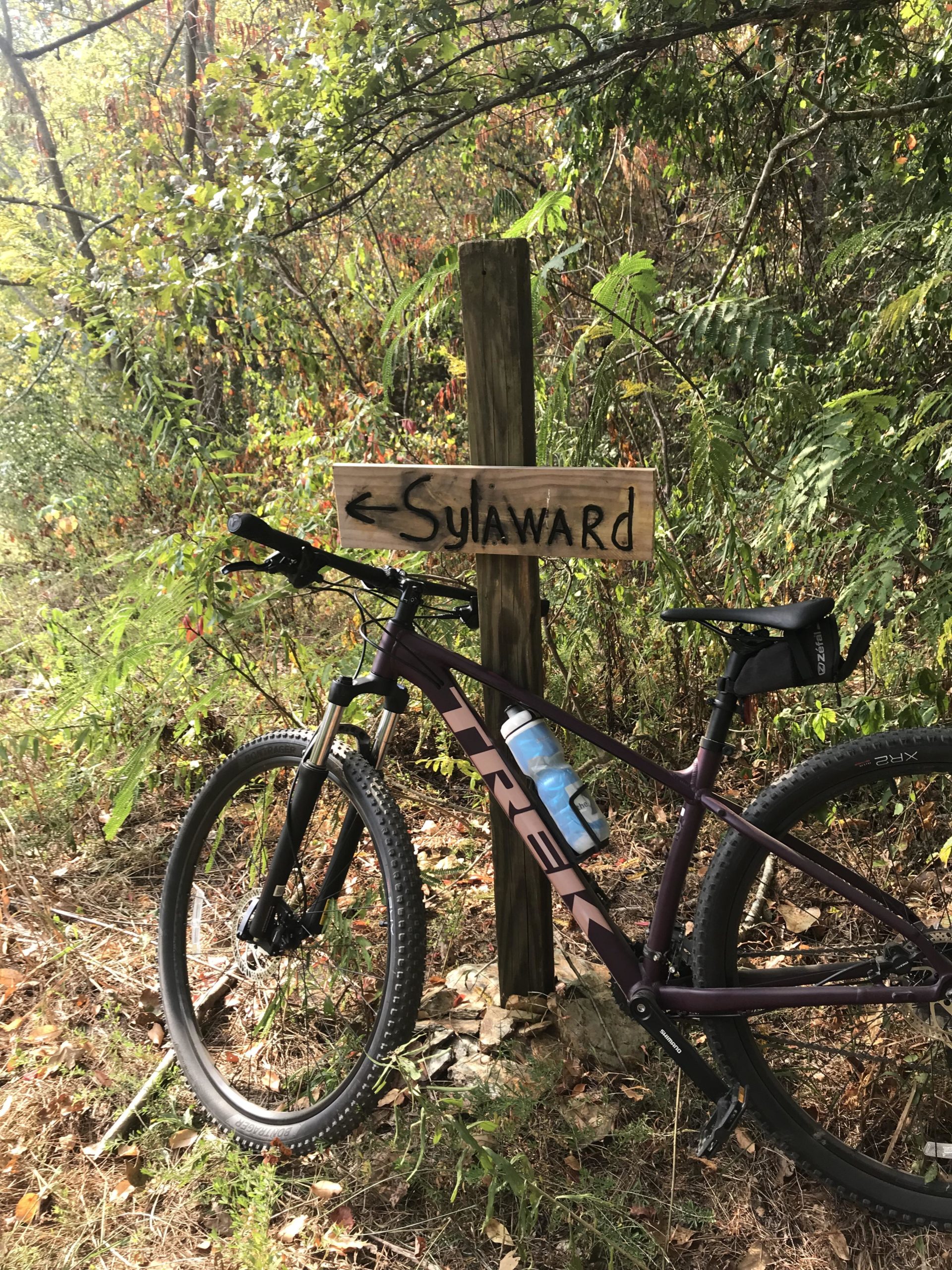 A mountain bike leaning against a wooden signpost labeled "Sylaward," surrounded by dense greenery and autumn foliage. Sylaward mountain bike trail.