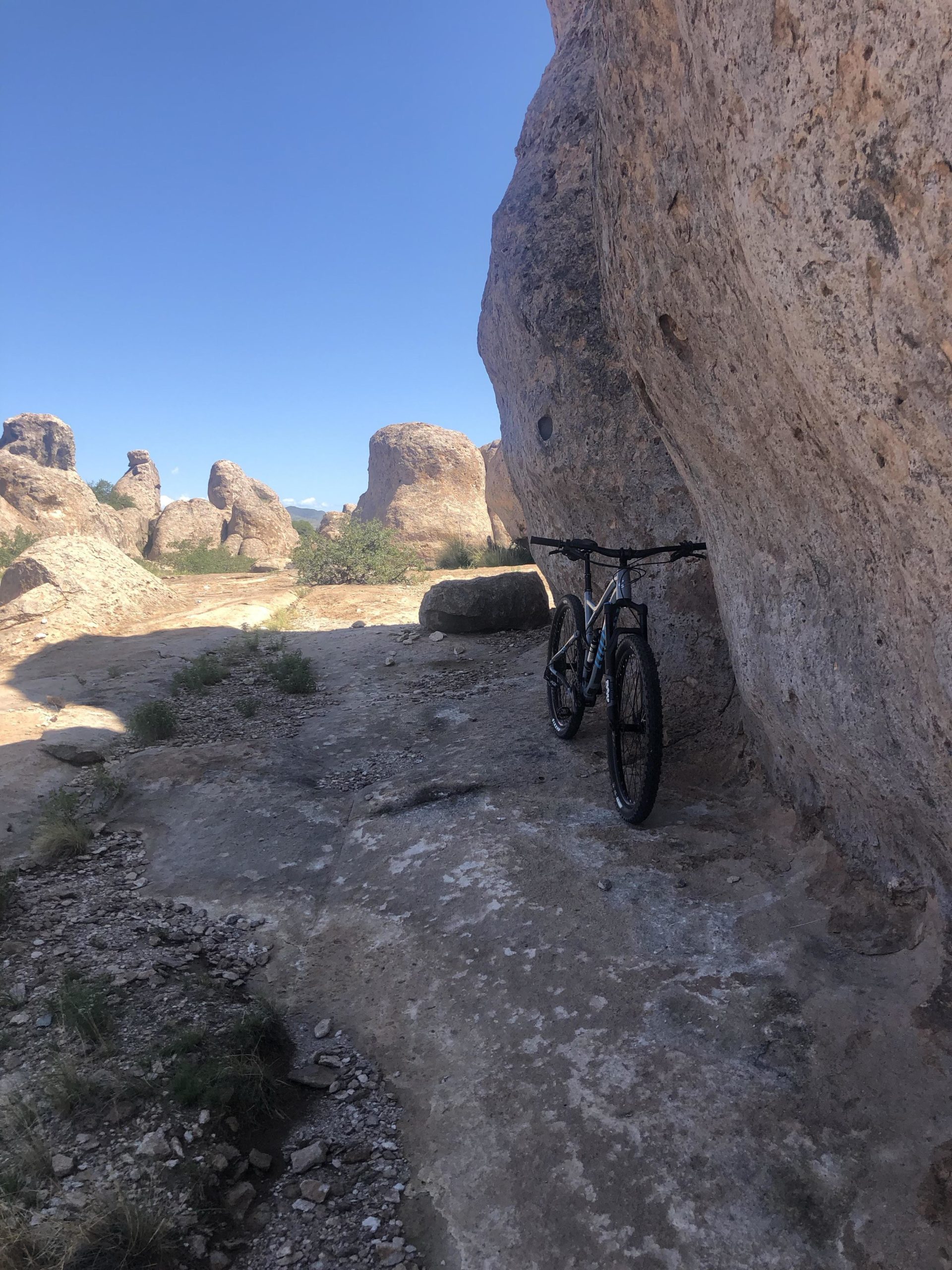 A mountain bike resting against a large rock, surrounded by rugged rock formations and a clear blue sky. The landscape features rocky terrain with sparse vegetation. City Of Rocks mountain bike trail.