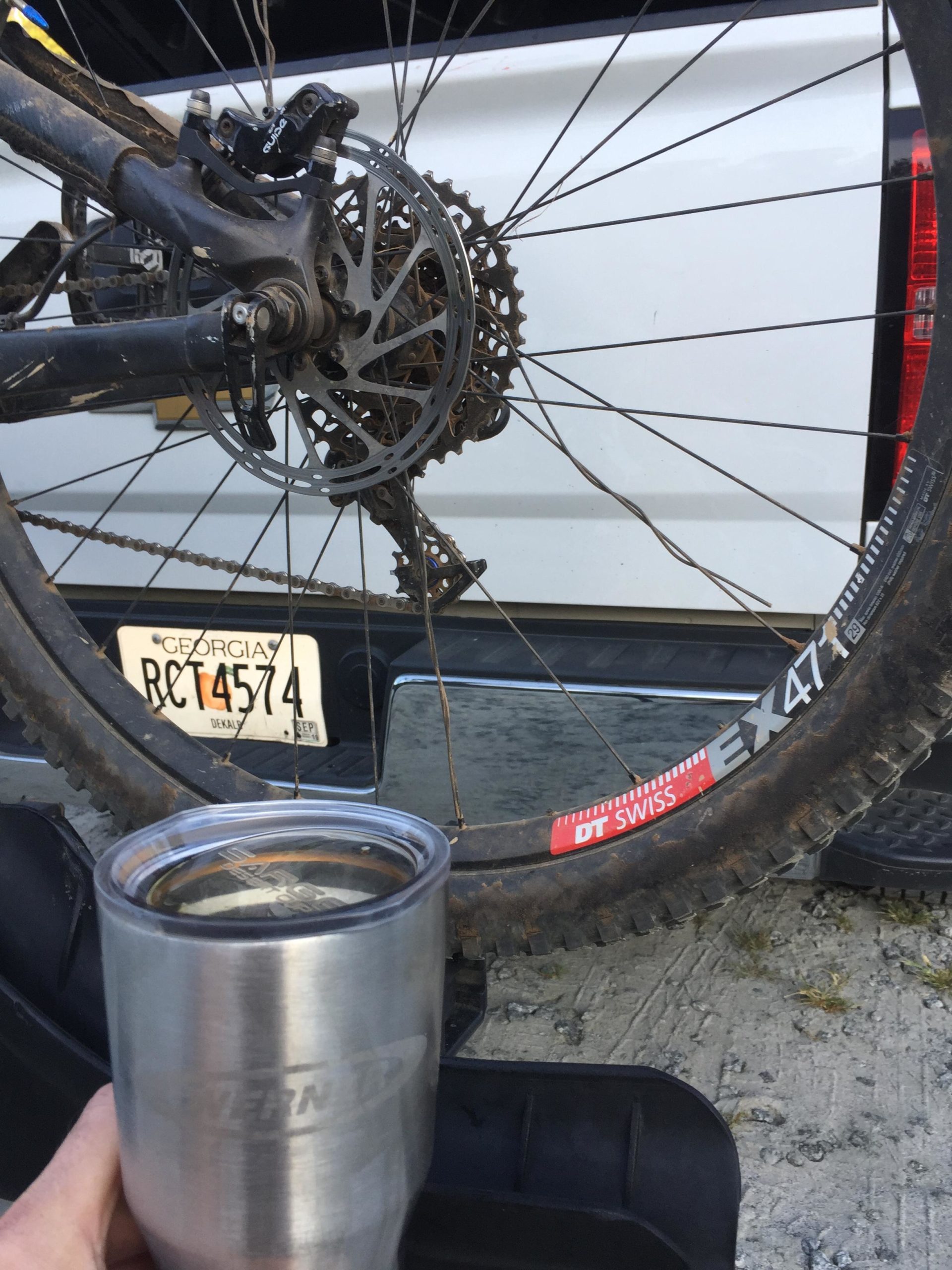 A close-up view of a bicycle's rear wheel and drivetrain, with a person's hand holding a metal drinking tumbler. The bicycle features a disc brake and a geared cassette, and the scene is set against the backdrop of a white truck and gravel terrain. Blankets Creek mountain bike trail.