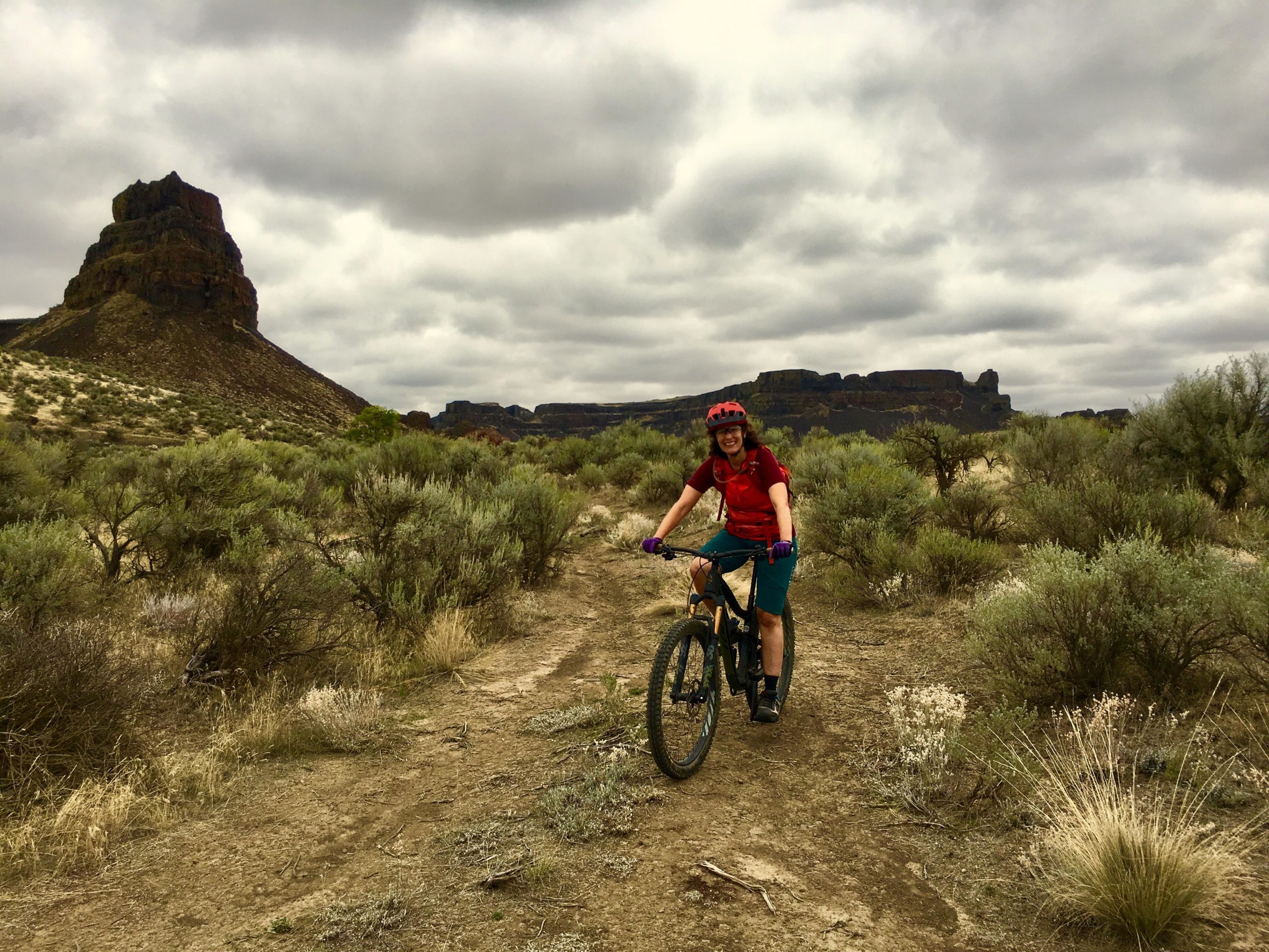 A person in a red shirt and helmet smiles while riding a mountain bike on a dirt path surrounded by sparse vegetation and rocky formations under a cloudy sky. Umatilla Trail mountain bike trail.