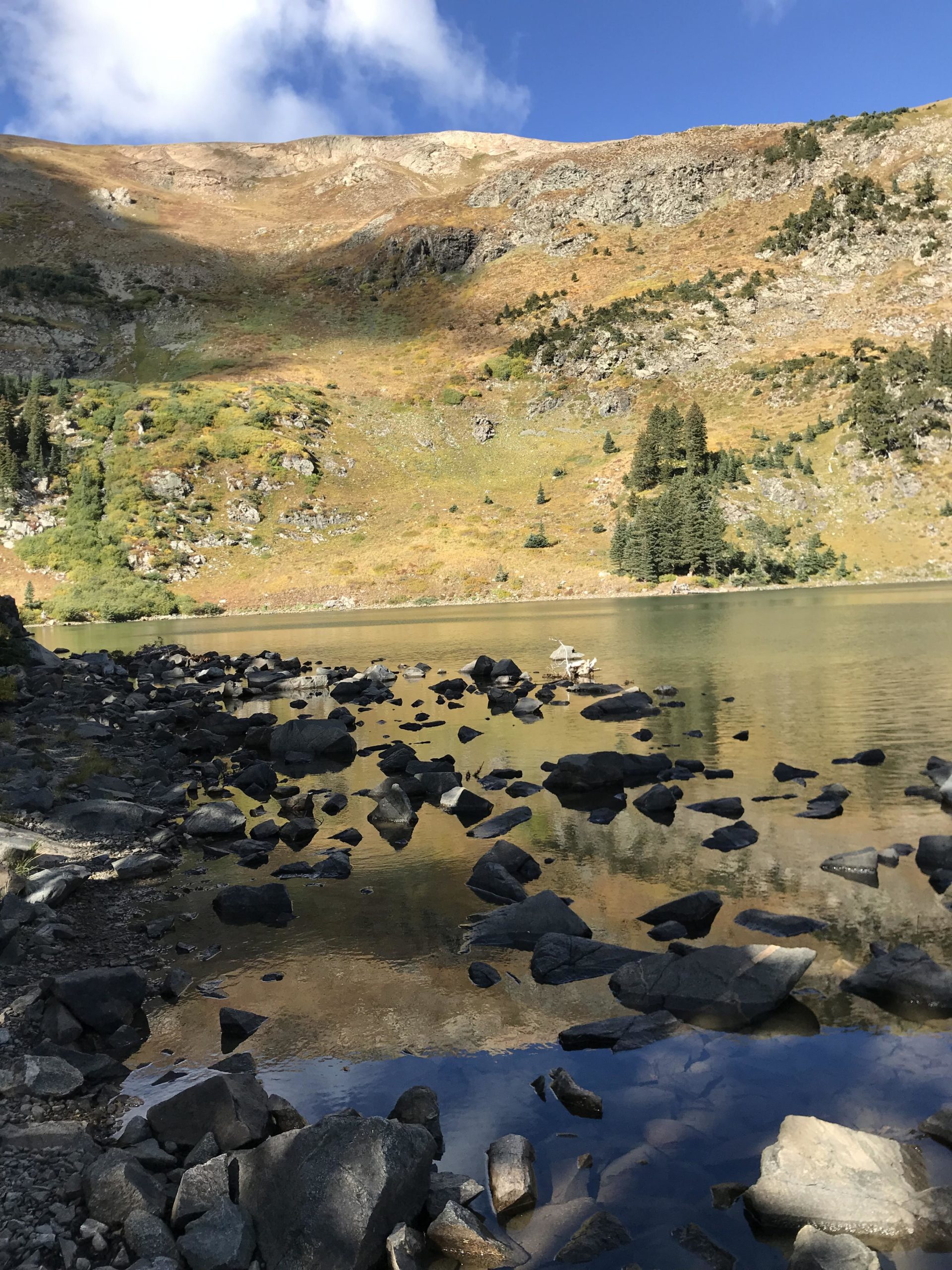 A serene mountain landscape featuring a calm lake surrounded by rocky shores. The hillside in the background is adorned with patches of greenery and rocky outcrops, under a clear blue sky with scattered clouds. The surface of the lake reflects the surrounding scenery, enhancing the tranquil atmosphere of the scene. Lost Lake to East Fork Loop mountain bike trail.