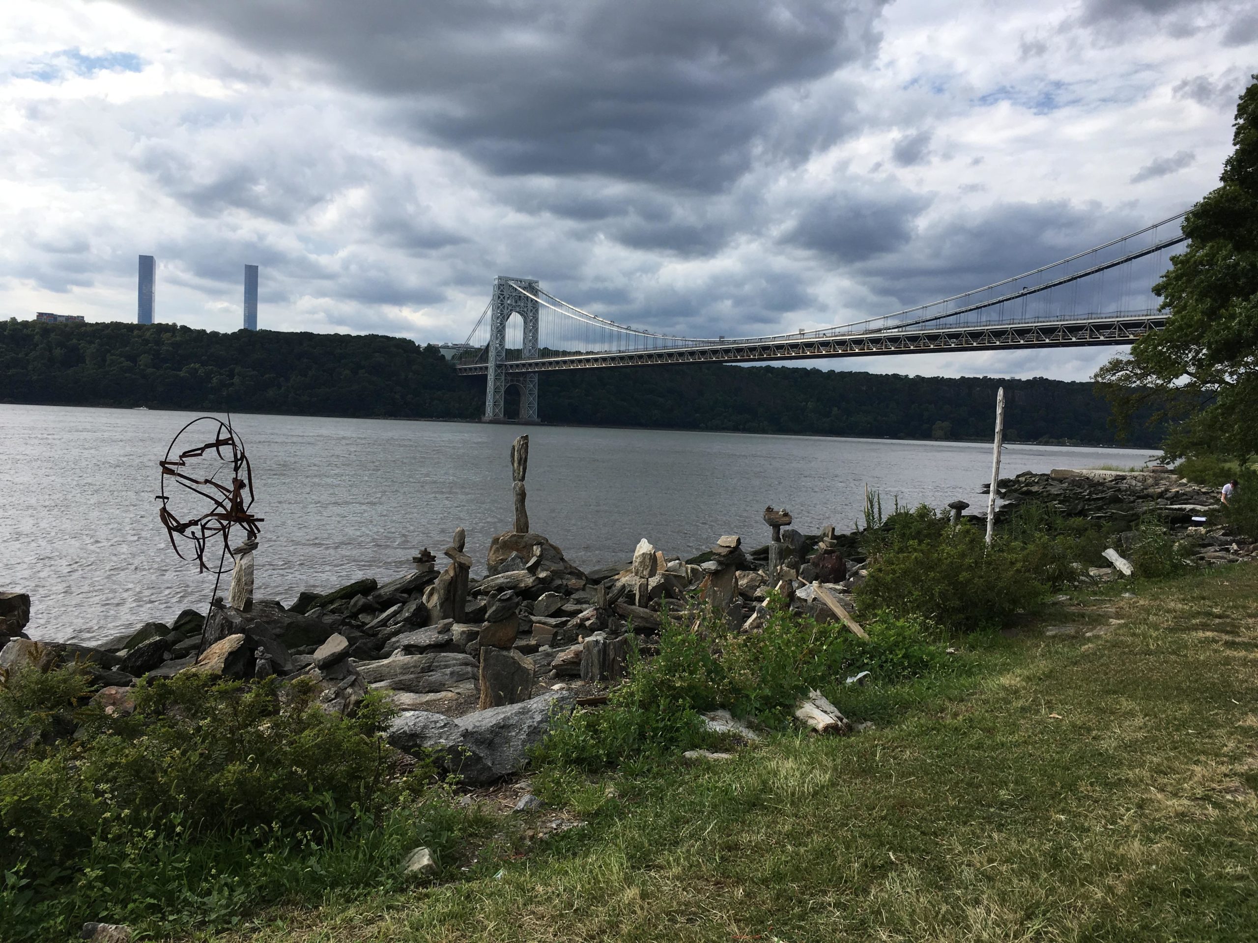 A view of the George Washington Bridge spanning the Hudson River, with a cloudy sky overhead. The foreground features a rocky shoreline and grassy areas, alongside a sculpture made from metal and wooden logs. In the background, two tall buildings are visible on the horizon, surrounded by lush greenery. West Street Greenway mountain bike trail.