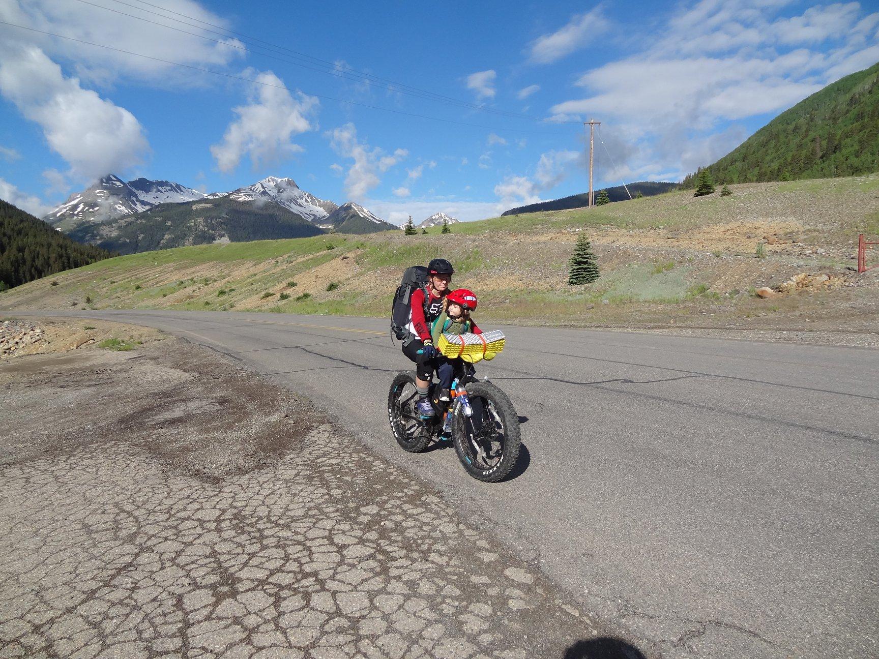 Salsa Mukluk: A cyclist riding a fat bike on a rural road, with a scenic mountainous landscape in the background. The cyclist is wearing a red jacket and a helmet, and there is a child seated comfortably in a basket on the bike. The sky is partly cloudy, and the terrain features greenery and rocky patches.