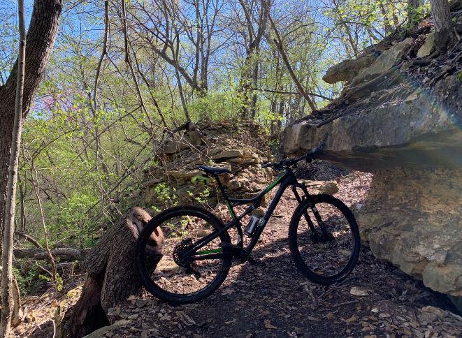 A mountain bike parked on a dirt trail surrounded by lush greenery and rocky formations under a clear blue sky. Big Bull Creek mountain bike trail.