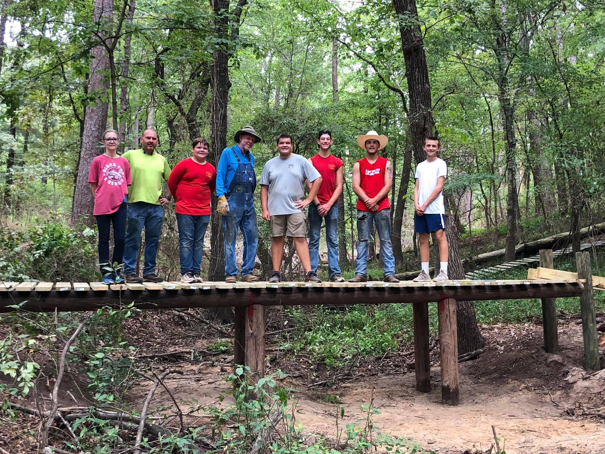 A group of nine people standing on a wooden bridge in a forested area. They are casually dressed and smiling, surrounded by trees and greenery. The scene conveys a sense of camaraderie and teamwork in a natural setting. Barber Hills Trail at Pat Mayse Lake mountain bike trail.